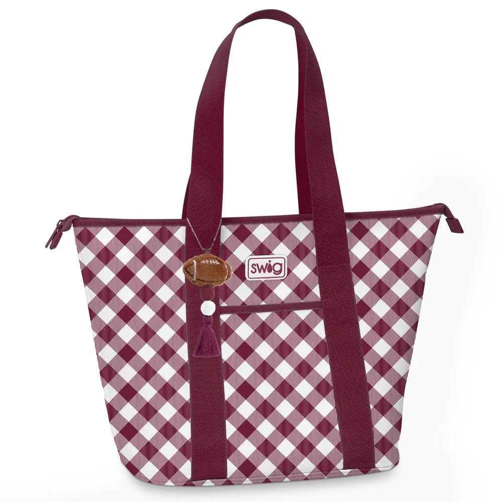 A Swig brand tote bag featuring a burgundy and white gingham pattern, with solid burgundy handles, a front zippered pocket, and a small brown football-shaped charm.
