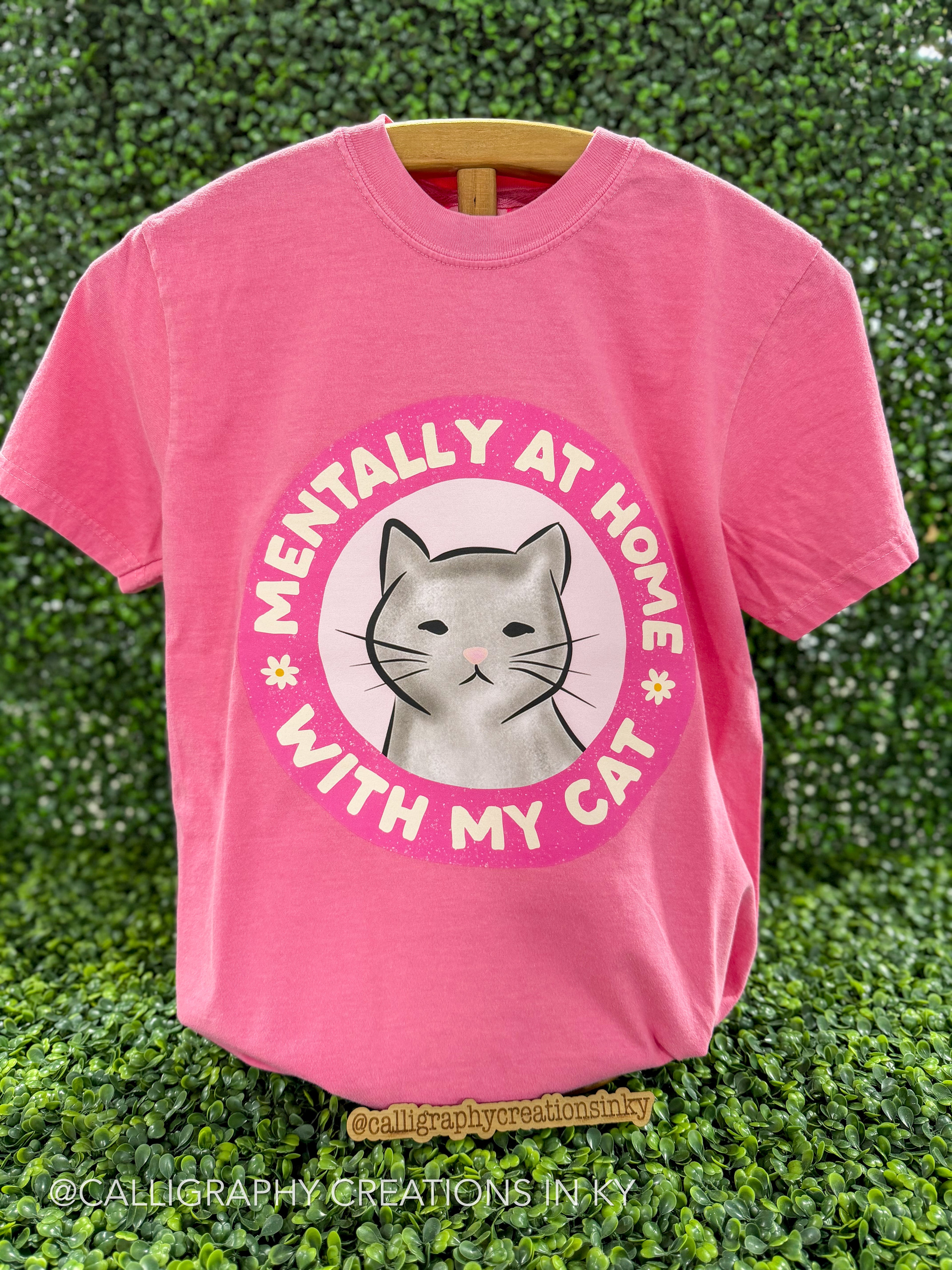 A pink short-sleeve t-shirt hanging on a wooden hanger against a green leaf wall. The shirt features a circular graphic with a gray cat illustration in the center and the text 'MENTALLY AT HOME WITH MY CAT' in pink and white.