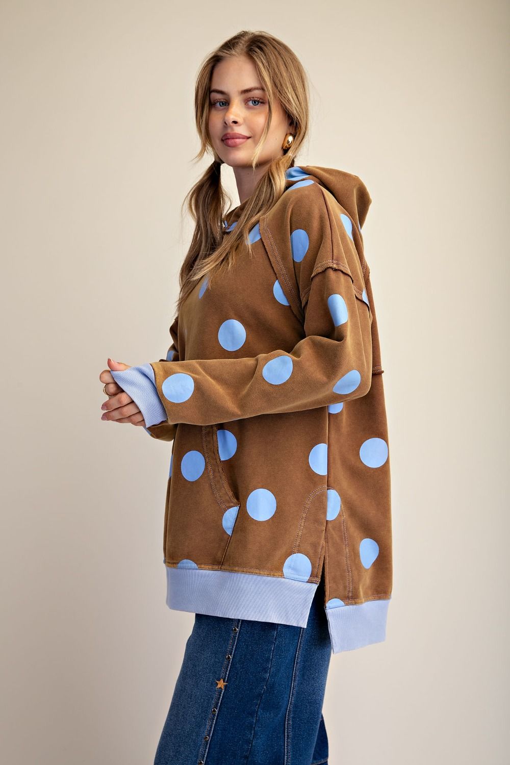 A woman modeling a brown hooded sweatshirt with large light blue polka dots and light blue ribbed cuffs and hem, paired with blue jeans against a plain beige studio background.