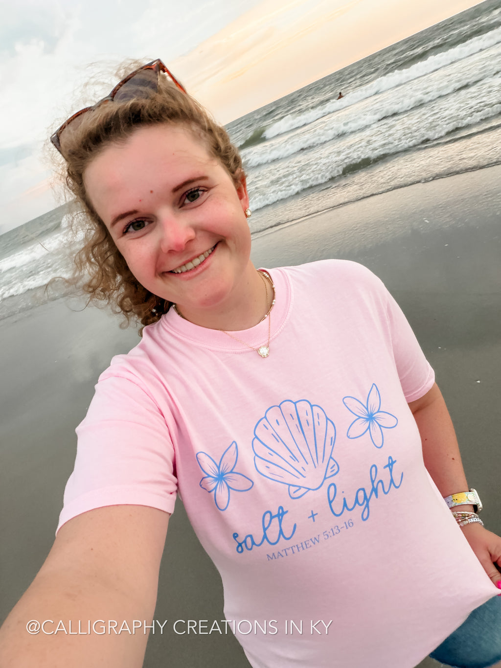A woman taking a selfie on a beach wearing a light pink t-shirt with a blue scallop and floral graphic and the text 'salt + light'.