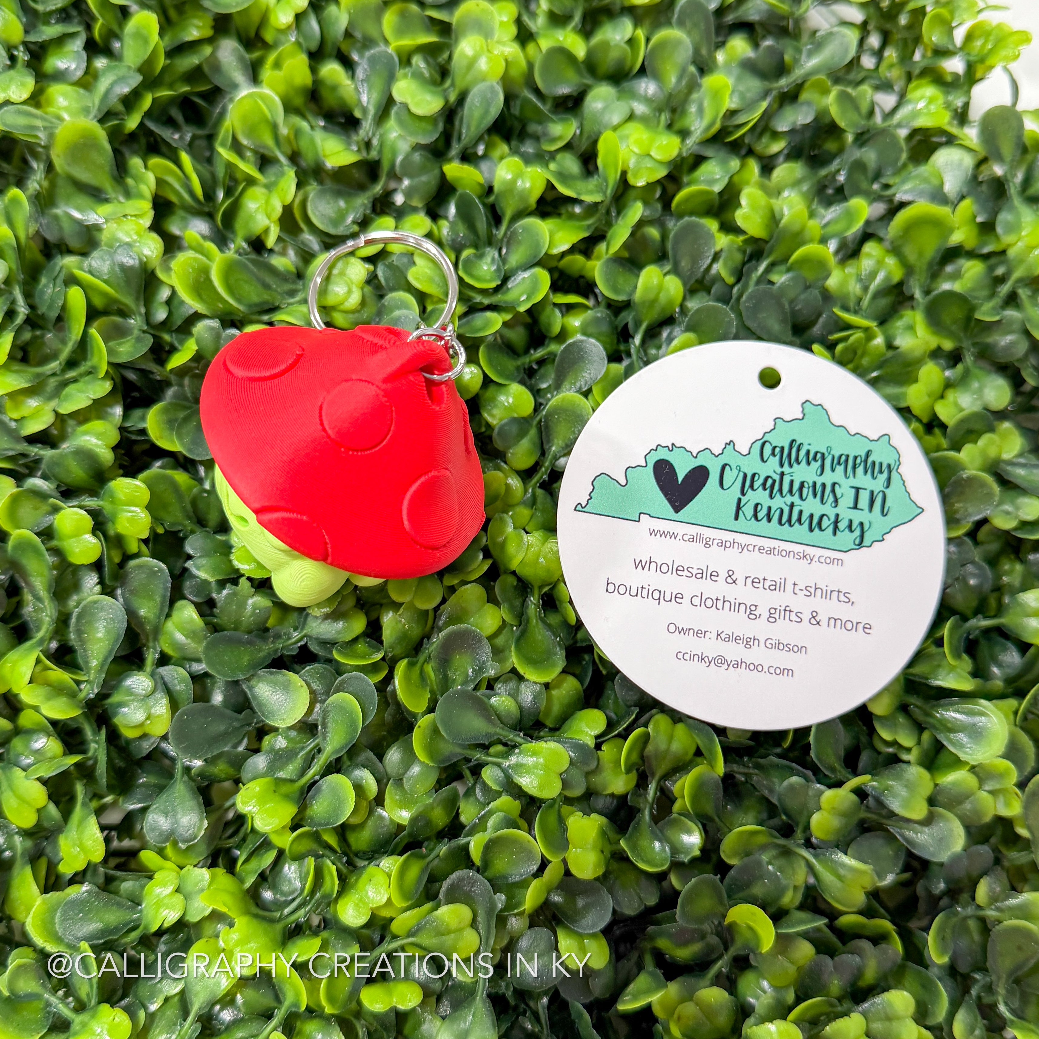 Side view of a Fidget clicker Keychain with a green frog with a red mushroom hat on a green leafy background with a Calligraphy Creations In Ky circle brand tag.