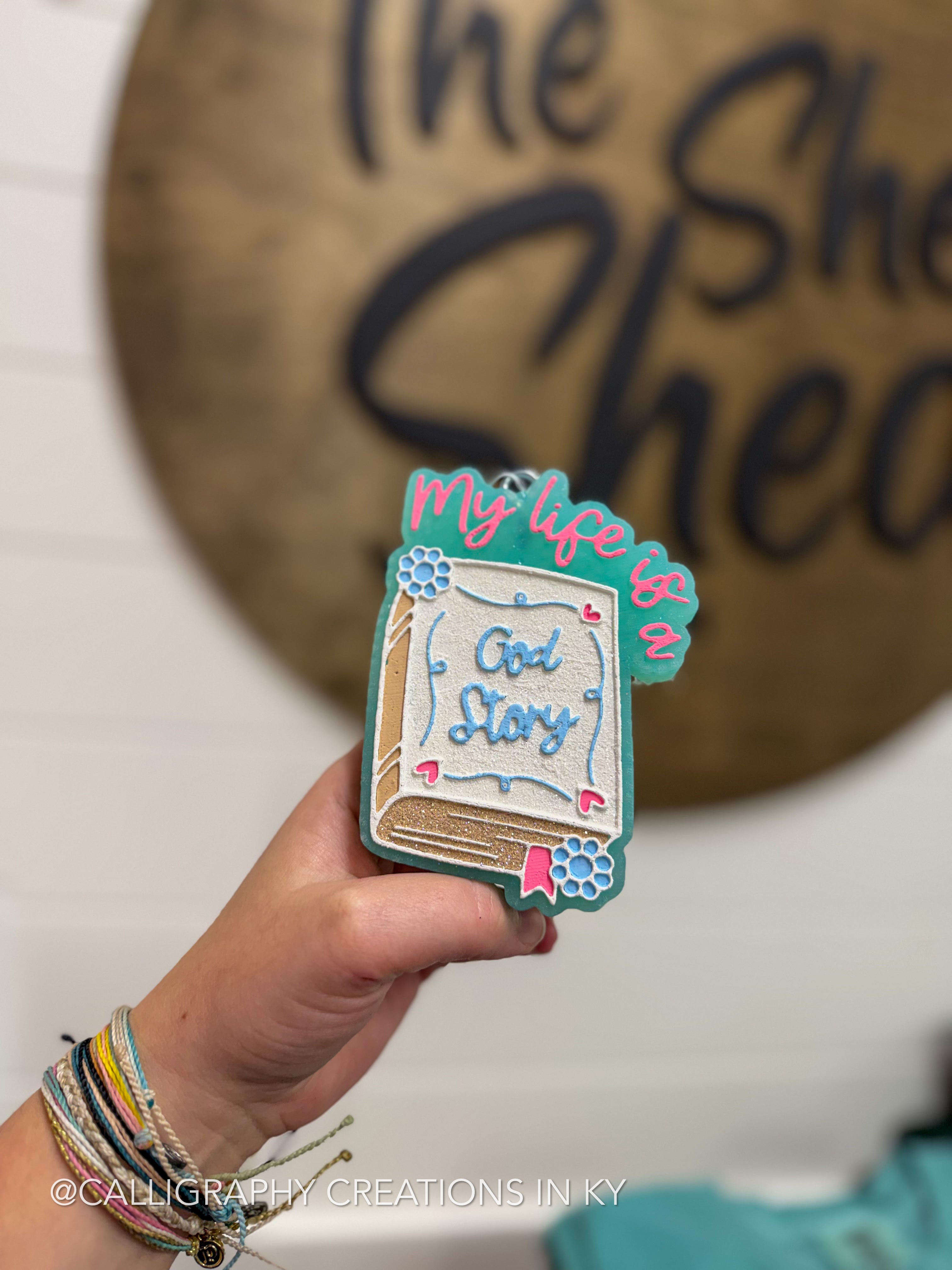 A hand holds a small decorative book-shaped item with the words 'My life is God Story' in blue and pink lettering, set against a blurred background with a wooden sign reading 'The She.