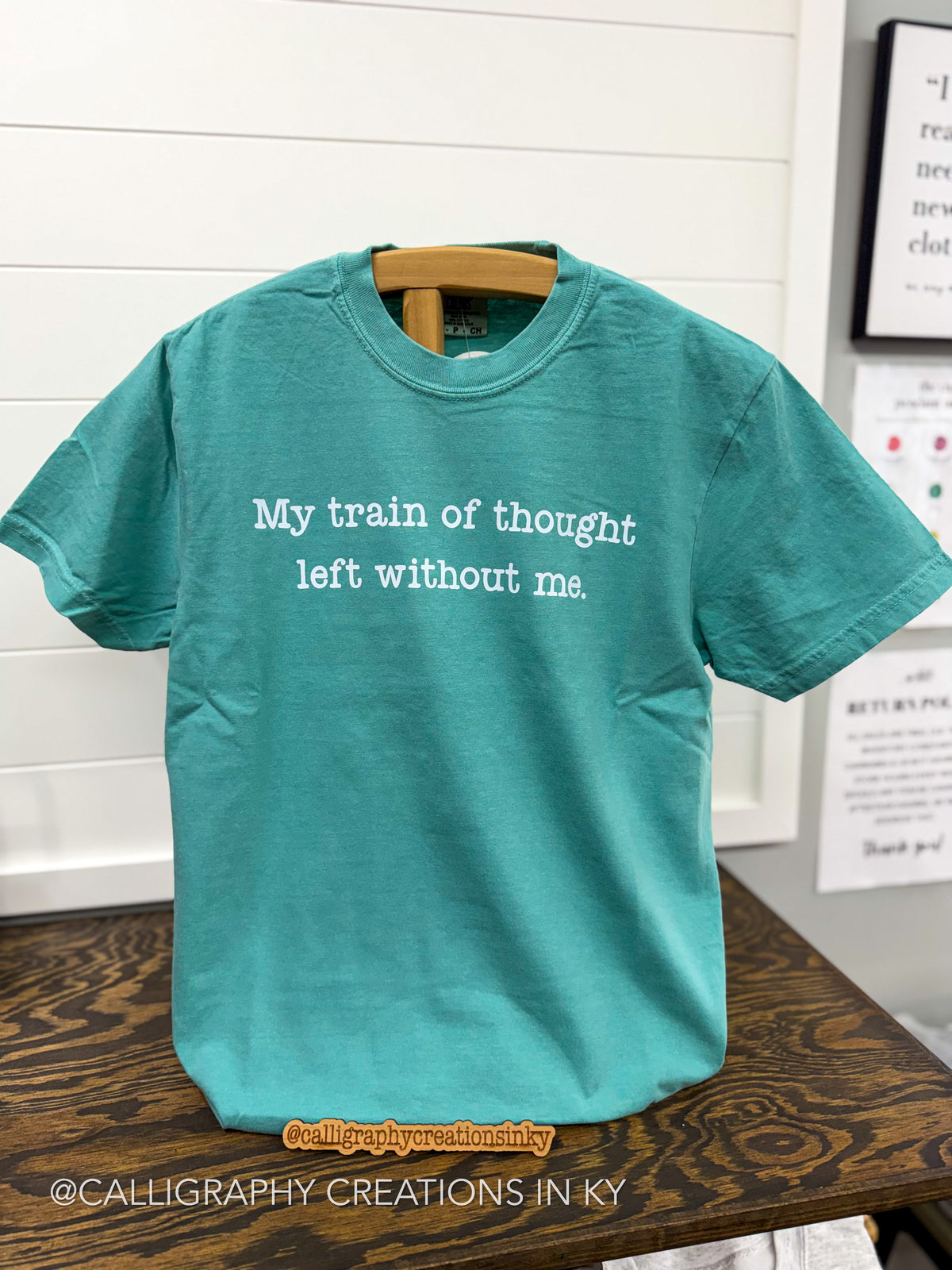 A teal short-sleeve t-shirt with white text reading 'My train of thought left without me.' hanging on a wooden hanger on a dark wood table against a white paneled wall.
