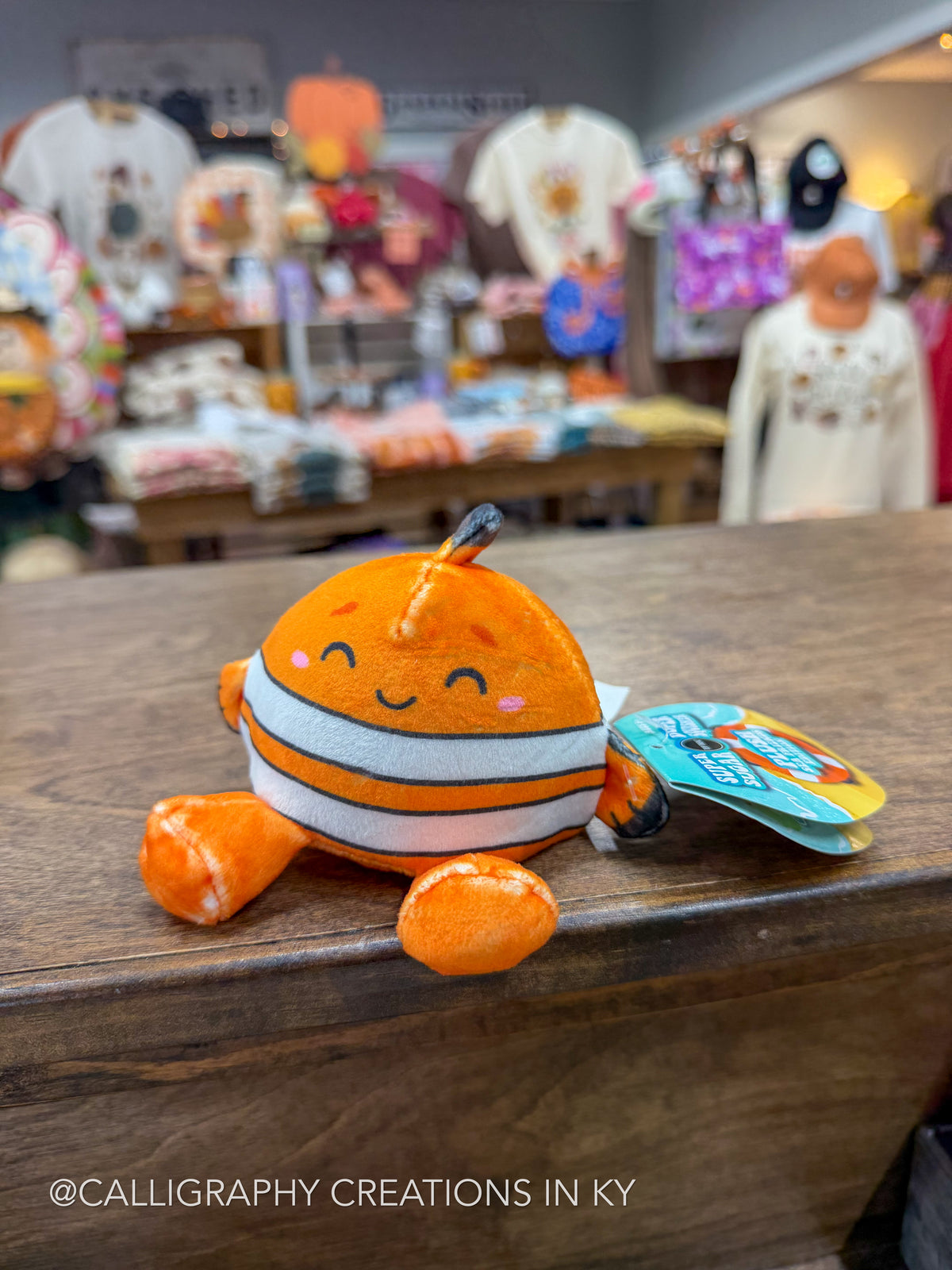 A small orange and white striped plush fish keychain with a smiling face, resting on a wooden table inside a retail store. A hangtag is attached to the side, and the background shows blurred merchandise and mannequins.