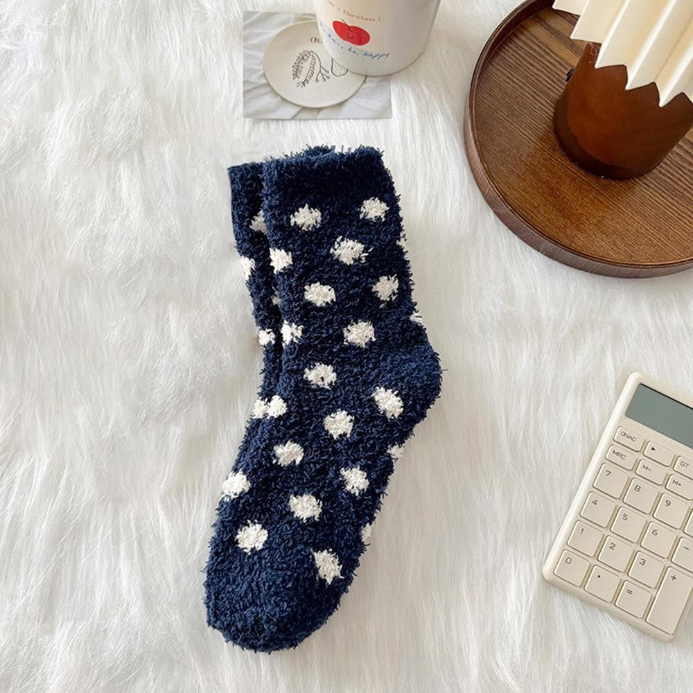 A single plush fuzzy sock in dark navy blue with white polka dots, laid flat on a white faux-fur surface with styled props including a wooden tray, cup, and calculator nearby.