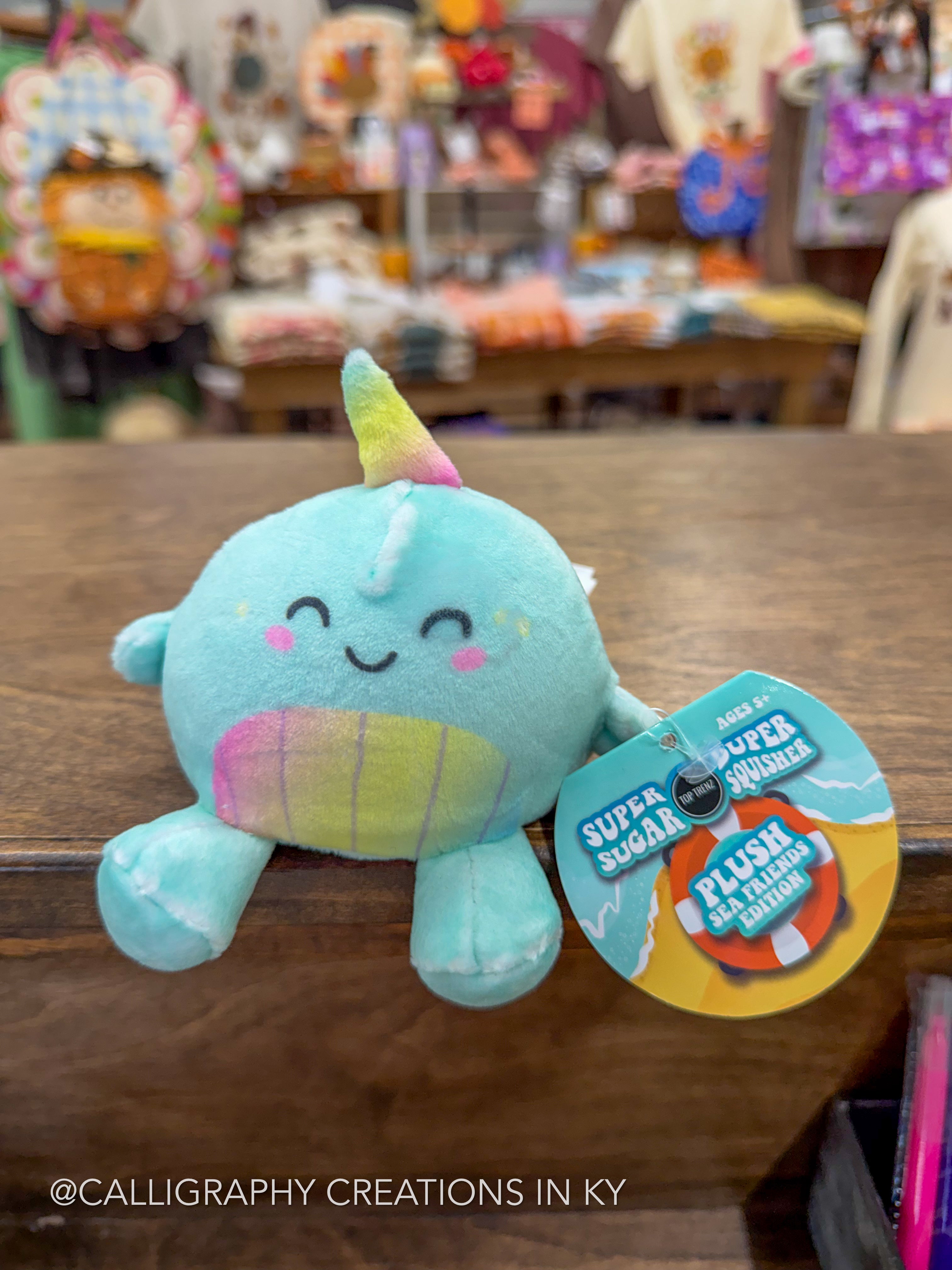 A light blue plush narwhal toy with a rainbow horn and a matching rainbow belly, sitting on a wooden table in a store. A product tag reading 'Super Sugar Plush Narwhal' is attached to its side.