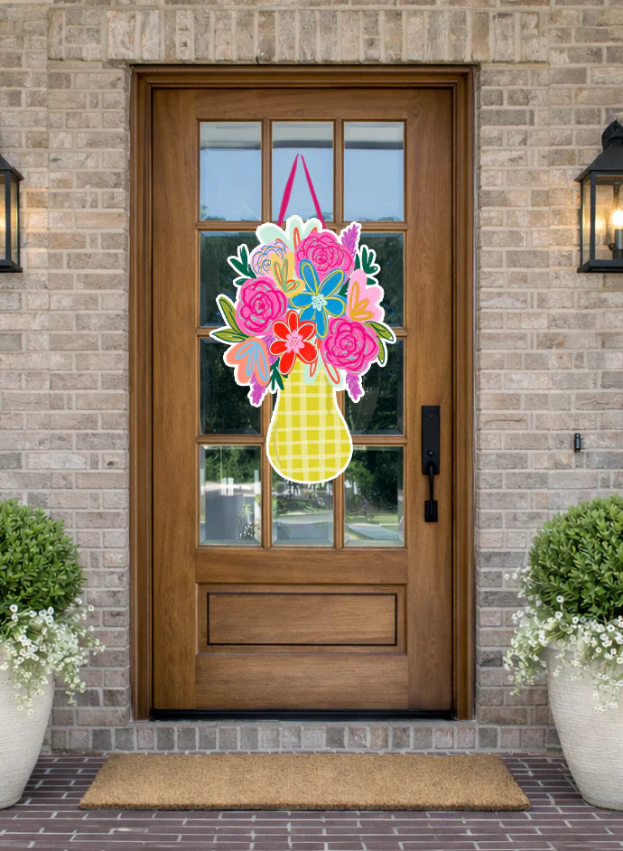 A colorful spring flower basket hanging on a brown wooden front door, flanked by potted plants and a brick wall.