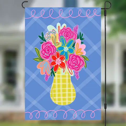 A decorative garden flag hanging from a black metal stand, featuring a colorful floral bouquet in a yellow checkered vase against a blue plaid background, with a blurred outdoor house and lawn behind it.