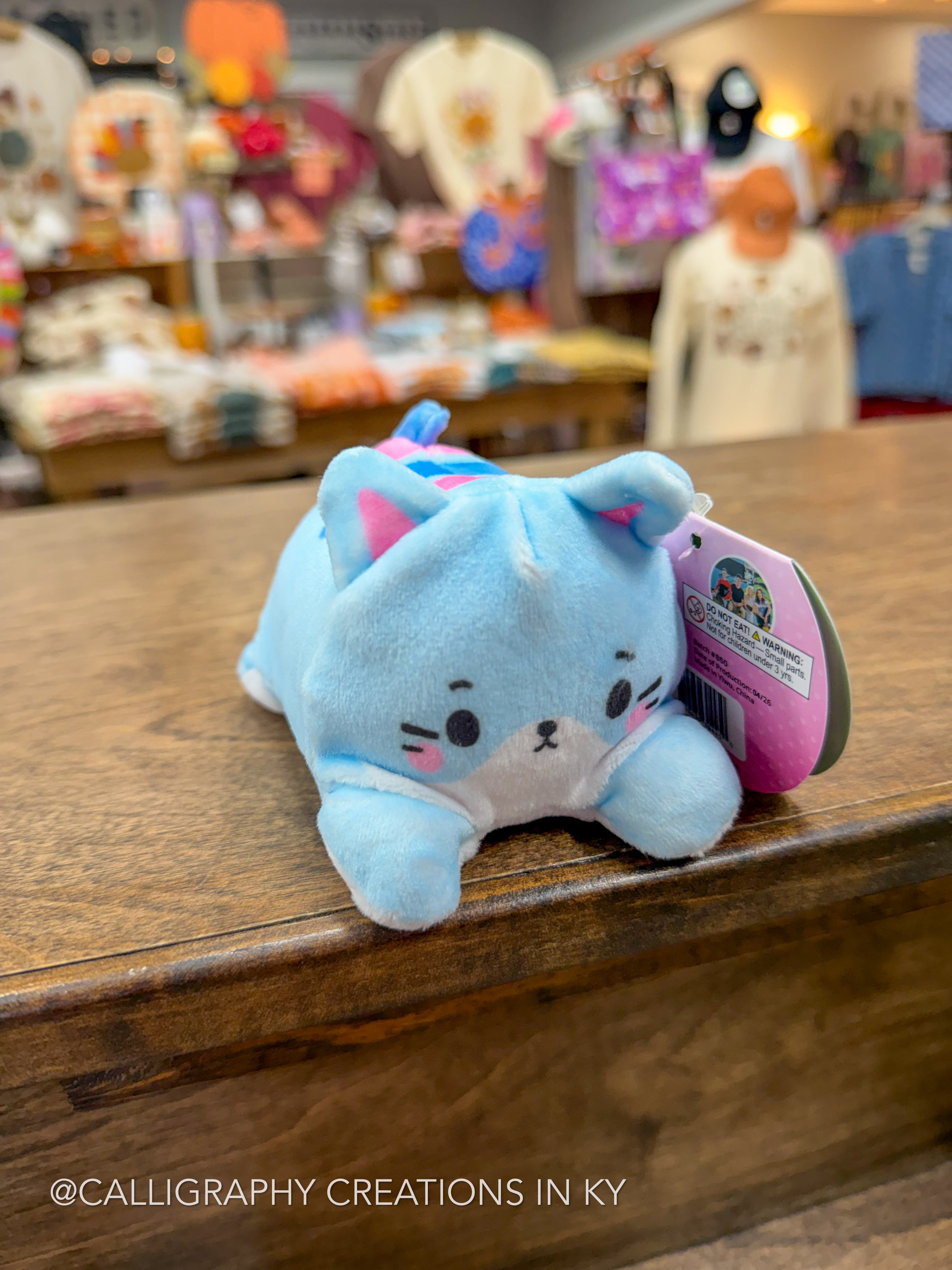 A light blue plush cat toy with pink and white accents, resting on a wooden counter inside a retail store. A pink and white hangtag is attached to the side of the plush.
