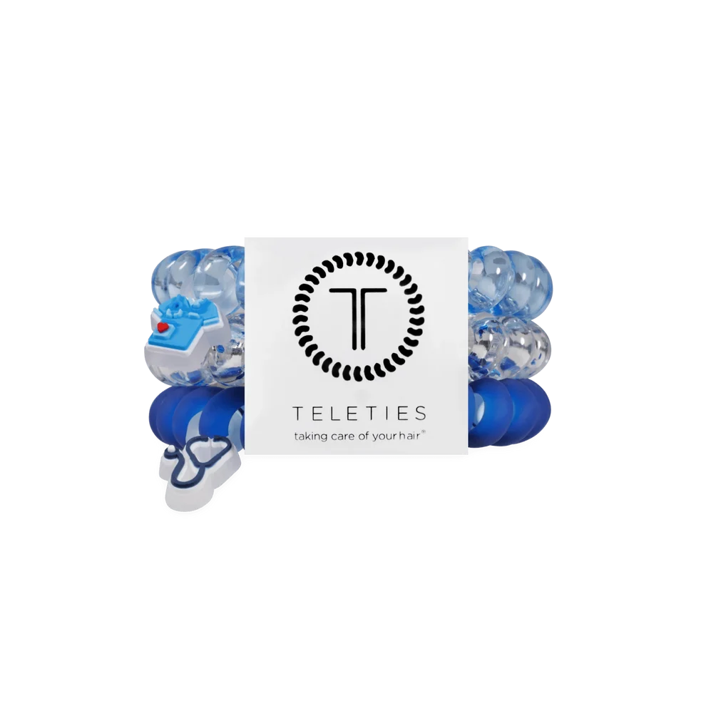 Three Teleties baby hair stretch bands in blue and clear, with a small blue and white heart-shaped charm, wrapped in a white paper band featuring the Teleties logo.