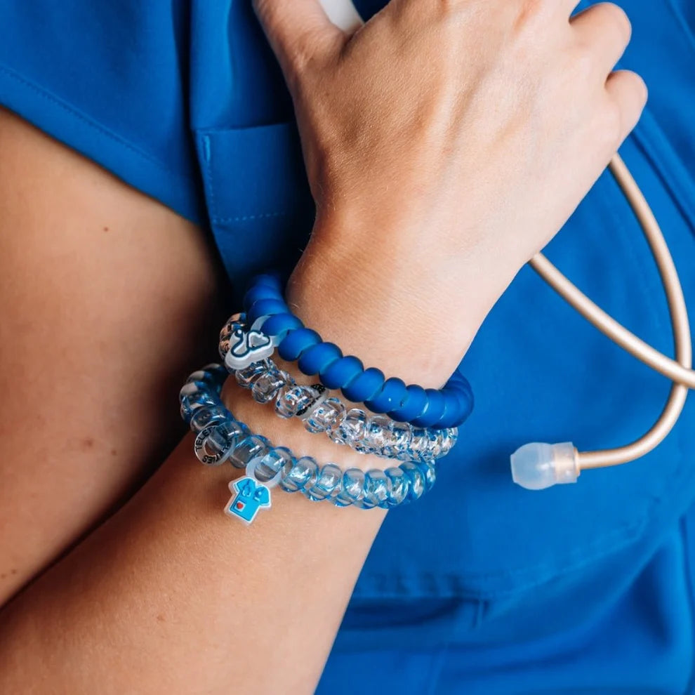 A close-up of a person's wrist wearing several blue beaded bracelets, including a blue silicone bracelet, a clear beaded bracelet with silver charms, and a clear beaded bracelet with a blue cross charm. The person is wearing blue medical scrubs and a stethoscope is visible.