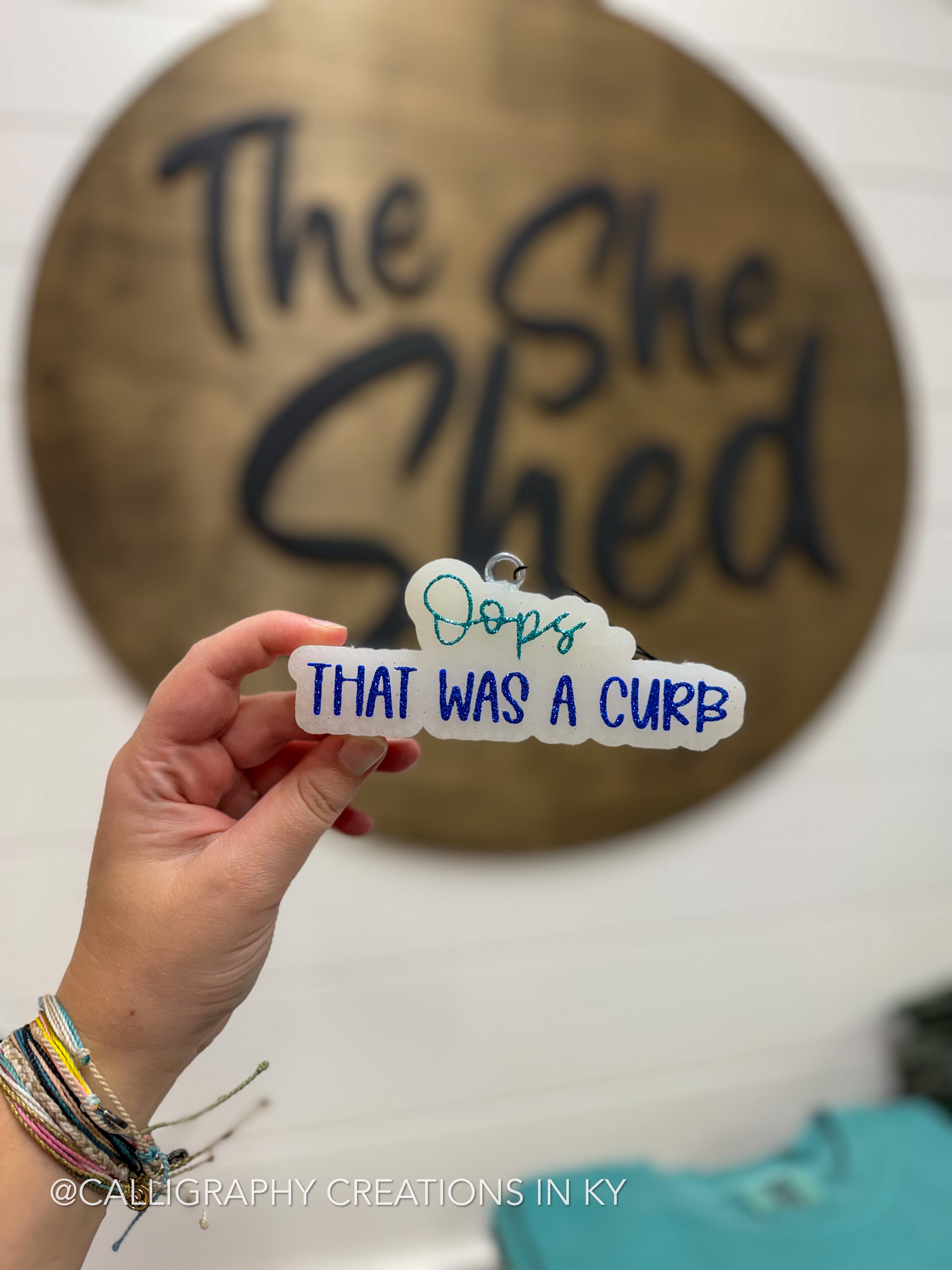 A hand holds a freshie that reads 'Oops THAT WAS A CURB' in blue and teal lettering on a white background. The sign is in front of a blurred wooden sign reading 'The She Shed' and a white wall.