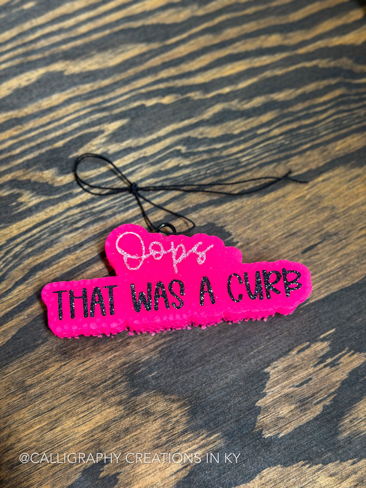 A bright pink calligraphic freshie reading 'Oops THAT WAS A CURB' in black and white, photographed on a dark wood surface. A black string is attached at the top, and a watermark at the bottom left reads '@CALLIGRAPHY_CREATIONS_IN_KY'.
