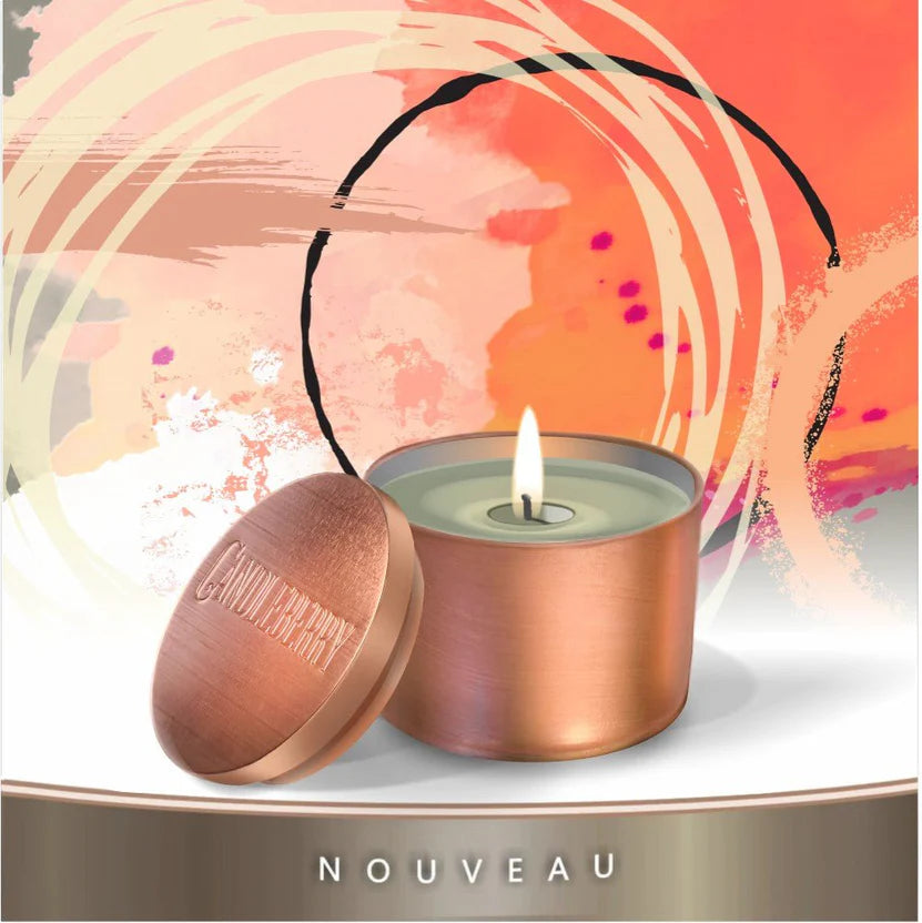 A lit candle inside a copper-colored travel tin with lid off to the side, displaying the word 'NOUVEAU' on a orange and pink swirled background.