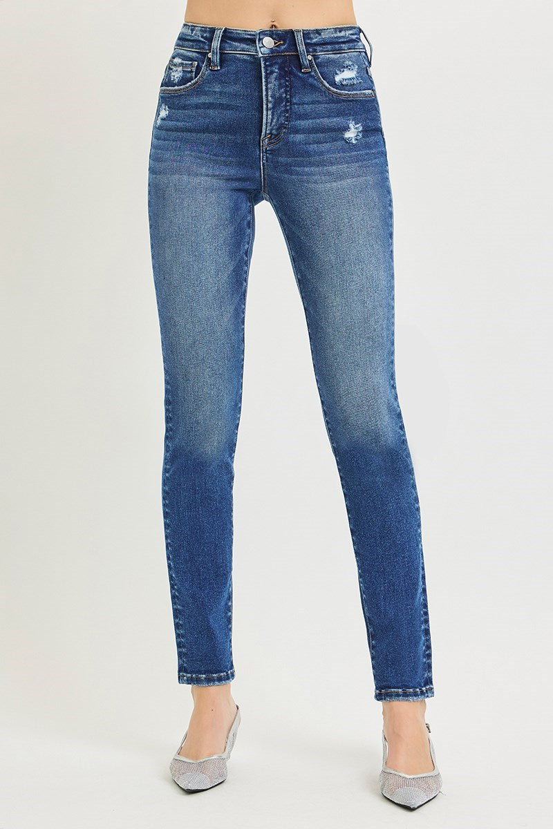 A person wearing medium-wash blue skinny jeans with distressed details on the thighs and knees, paired with pointed silver heels, shown against a plain light gray studio background.