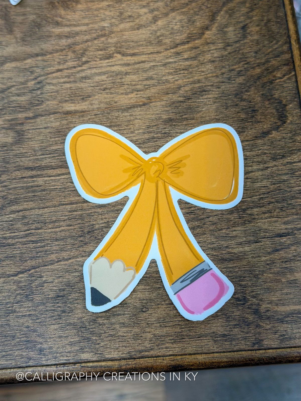 A die-cut sticker shaped like a yellow bow made from two crossed pencils, placed on a dark wood surface. The sticker has a white border and small pink and beige eraser tips.