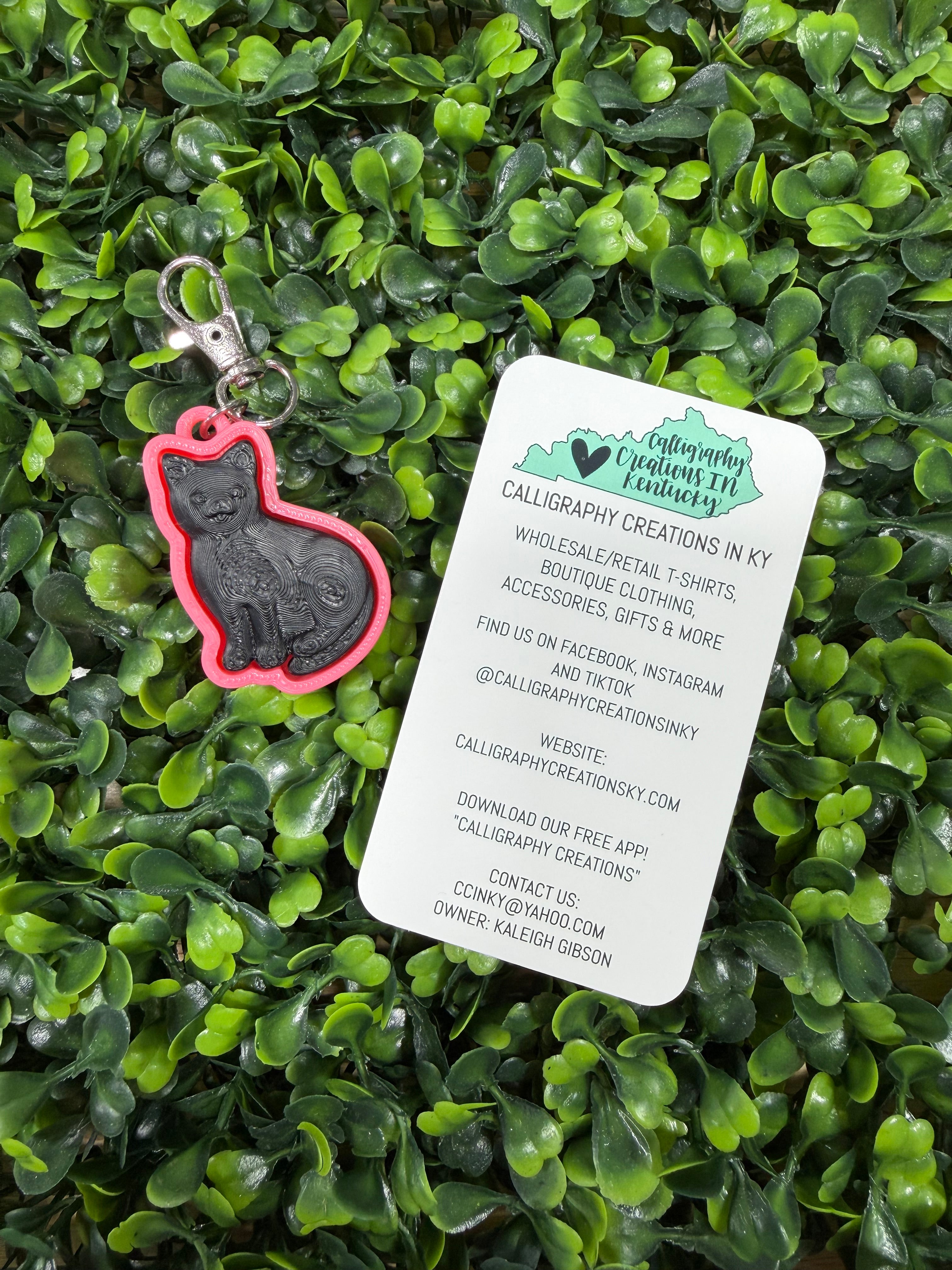 A pink and black cat-shaped fidget keychain with a silver clasp is displayed on a bed of green boxwood leaves next to a white business card for Calligraphy Creations by Kalder Gibson.