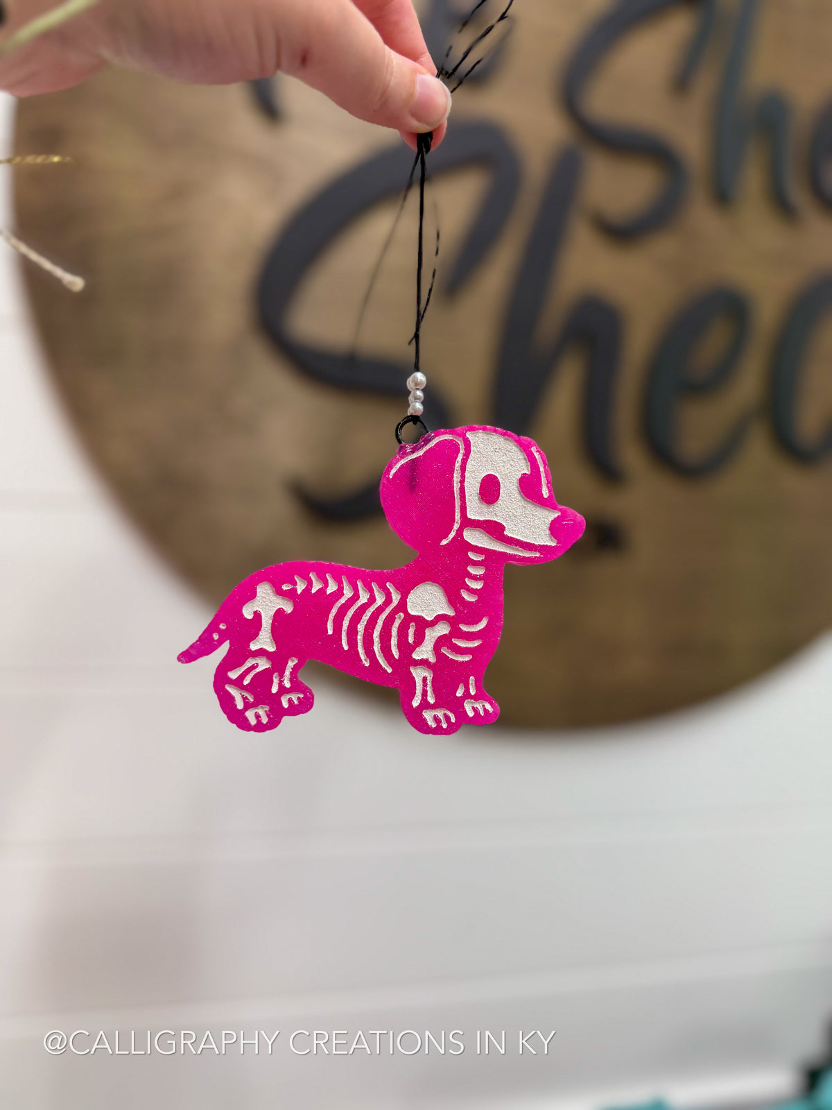 A hand holds a bright pink dachshund-shaped ornament with a white skeleton pattern, featuring a black hanging string against a blurred background with a wooden sign.