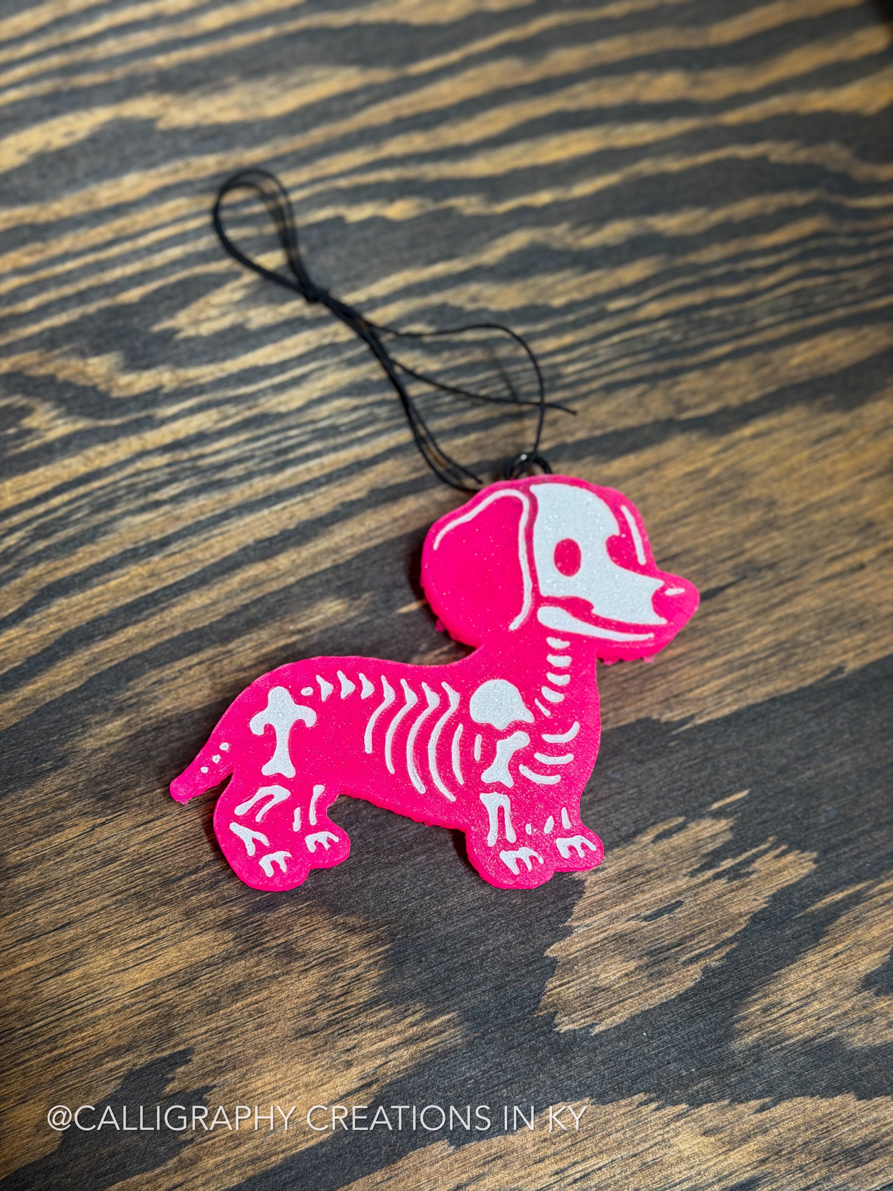 A bright pink dachshund-shaped ornament or charm with a white skeleton pattern and a black hanging loop, placed on a dark wood surface.