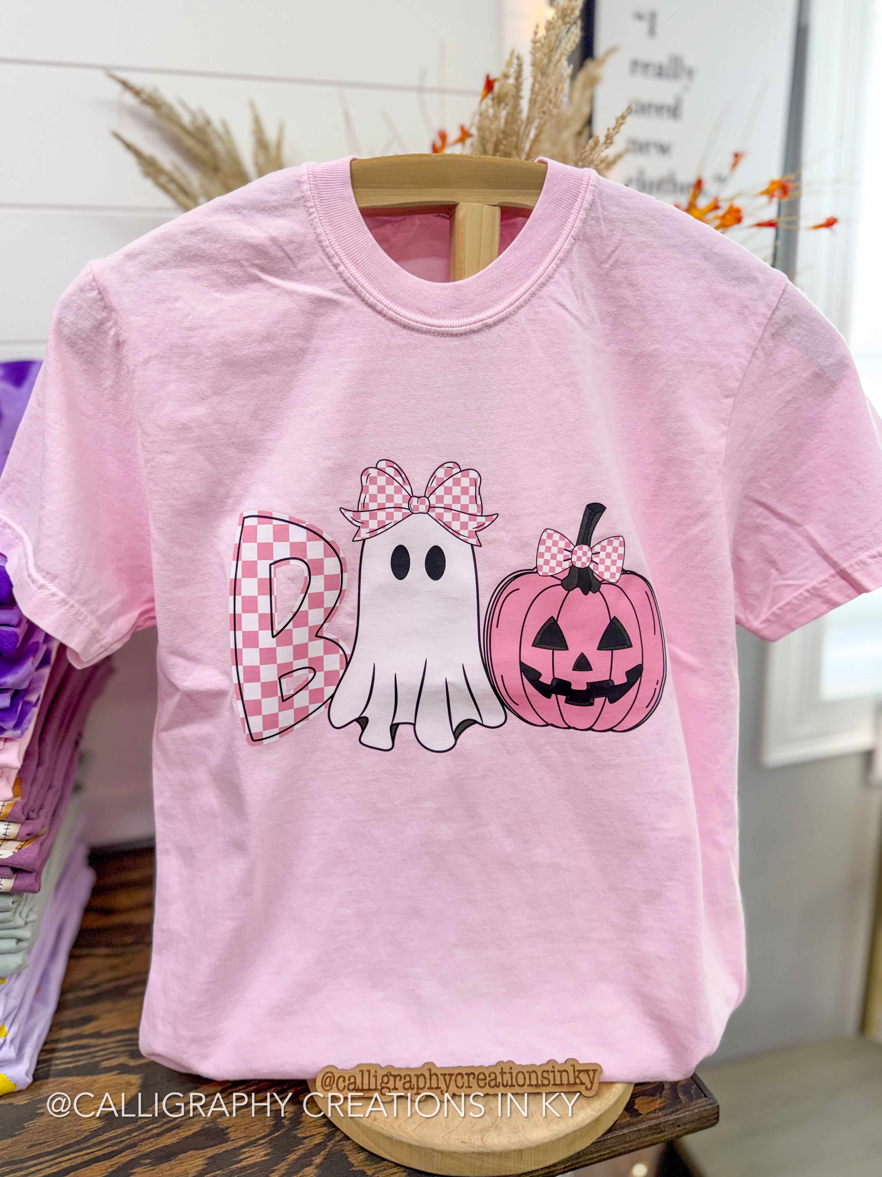 A light pink short-sleeve t-shirt displayed on a wooden hanger in a retail setting. The front features a Halloween-themed graphic spelling 'Boo' with a pink-and-white checkered 'B', a white ghost with a pink-and-white checkered bow, and a pink jack-o'-lantern pumpkin with a matching bow.