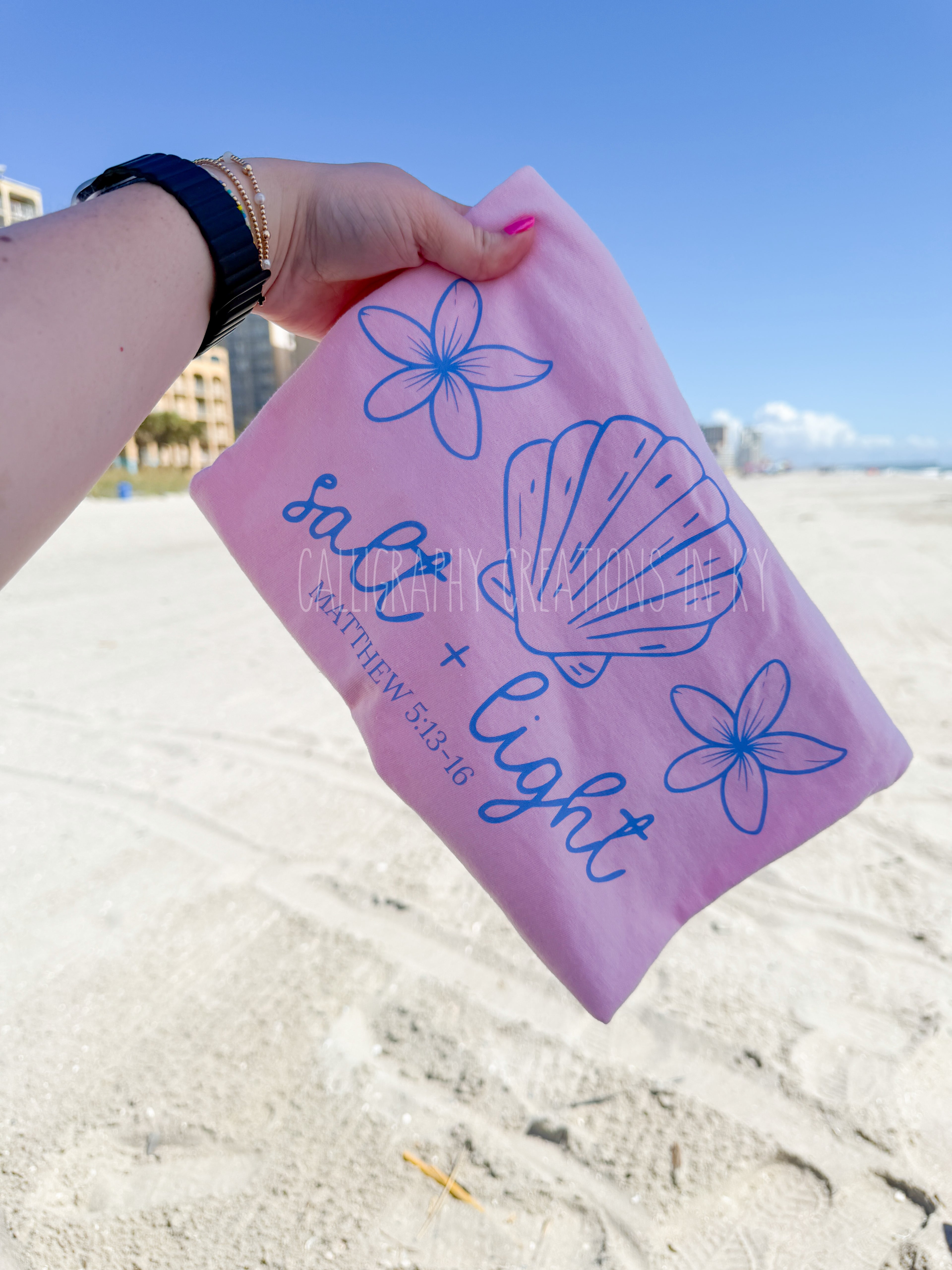 A hand holding a pink t shirt with blue line drawings of flowers and a seashell, with the text 'salt + light' and 'MATTHEW 5:13-16' printed on it, set against a sandy beach background.