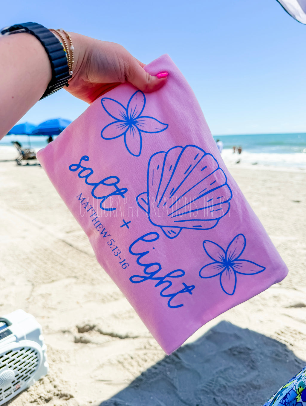 A hand holding a pink t shirt with blue line drawings of flowers and a seashell, with the text 'salt + light' and 'MATTHEW 5:13-16' printed on it, set against a sandy beach background.