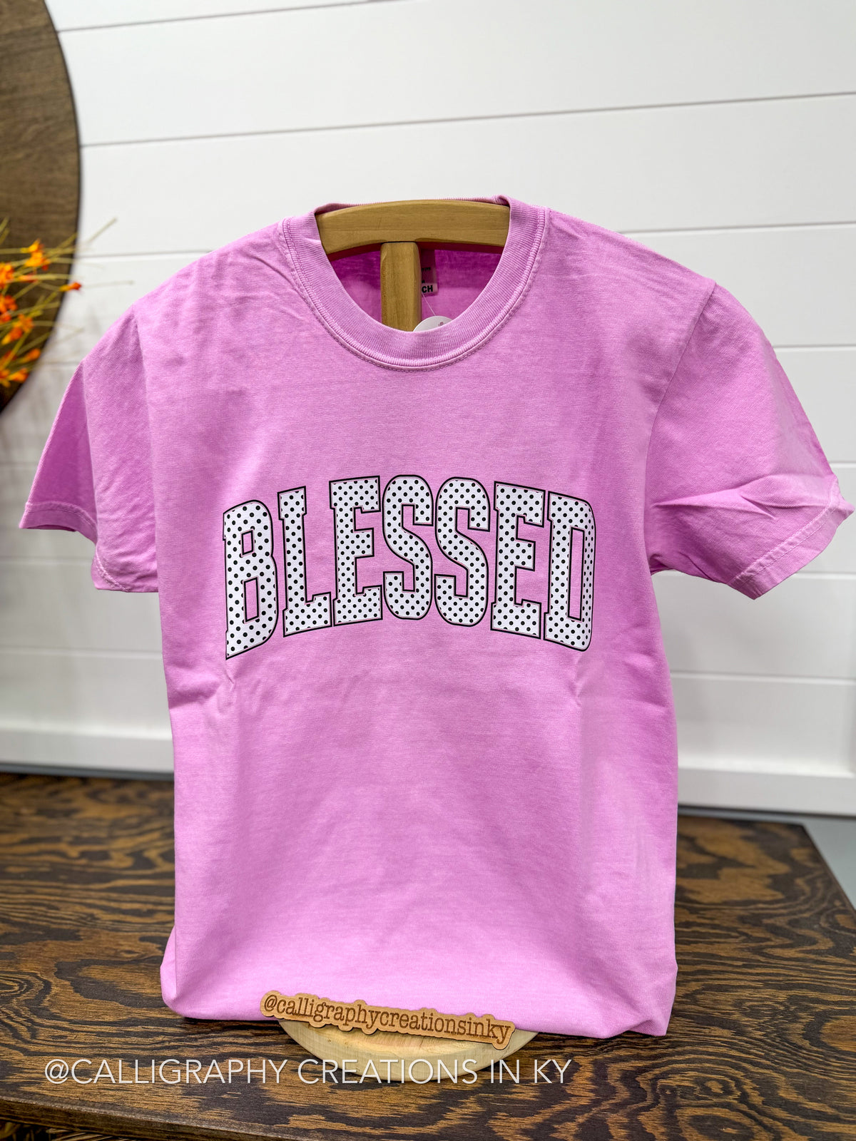 A pink short-sleeve t-shirt with 'BLESSED' printed across the chest in white letters filled with black polka dots. The shirt is displayed on a wooden hanger on a dark wood table against a white shiplap wall.