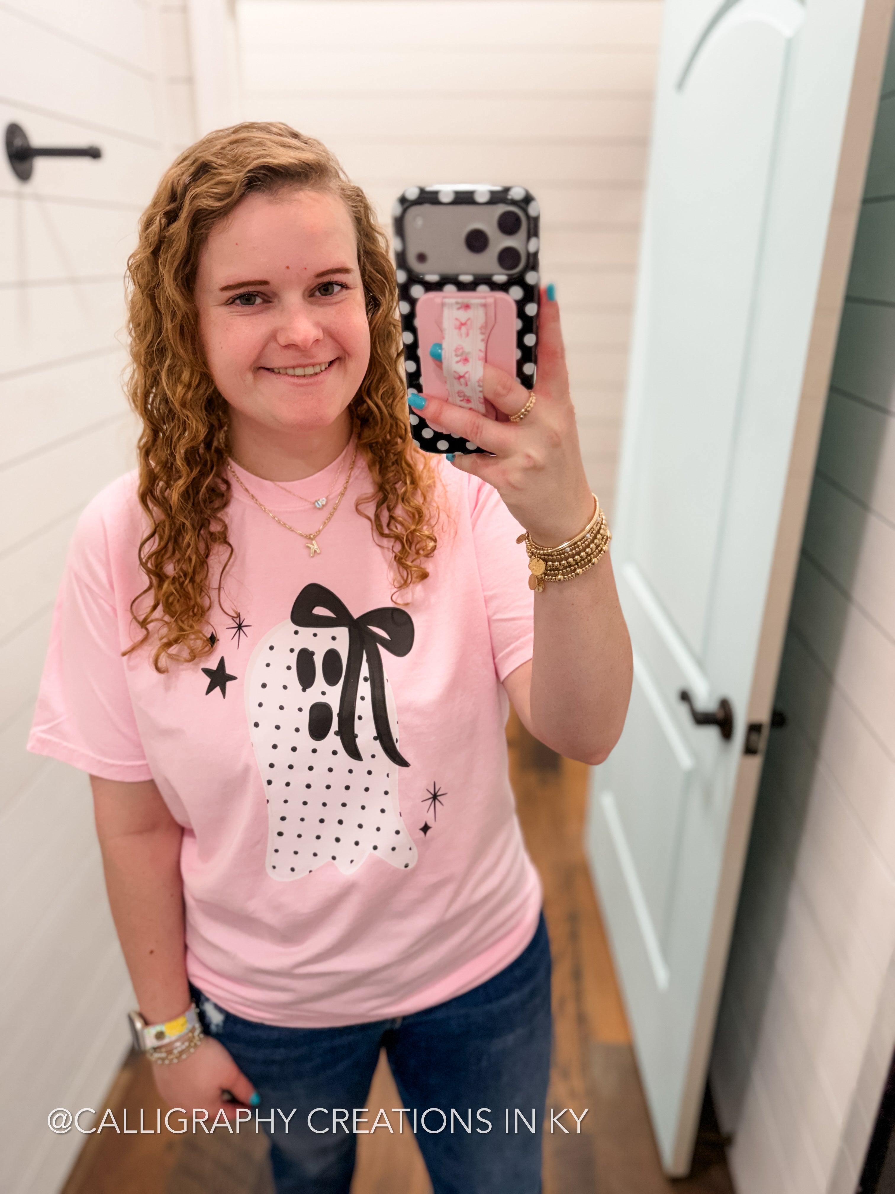 A woman takes a mirror selfie wearing a light pink t-shirt with a black and white polka dot bow graphic, set in a bright room with white paneled walls and doors.