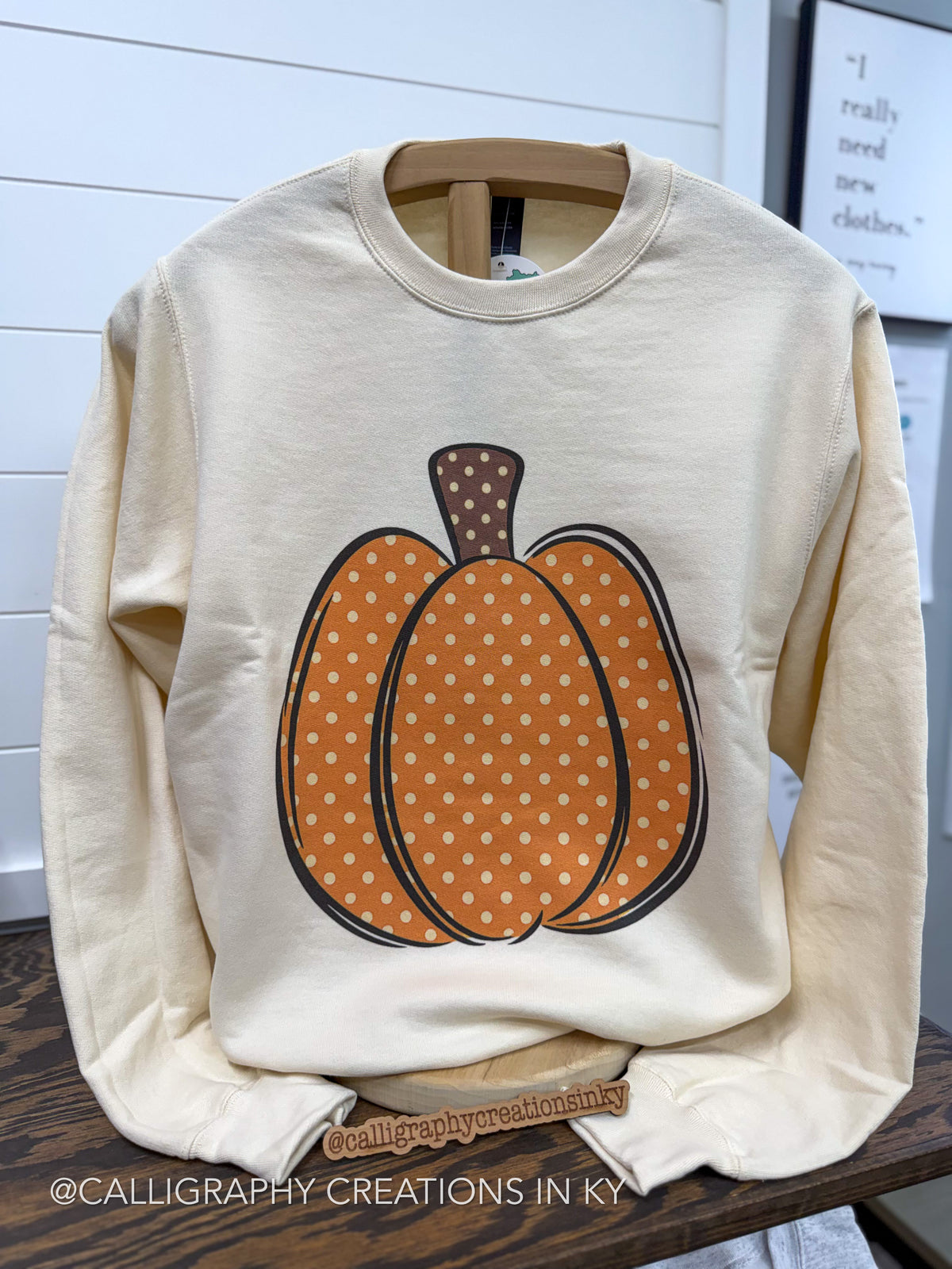 A cream-colored crewneck sweatshirt featuring a large orange pumpkin graphic with white polka dots and a brown stem, displayed on a wooden hanger on a dark wood table.