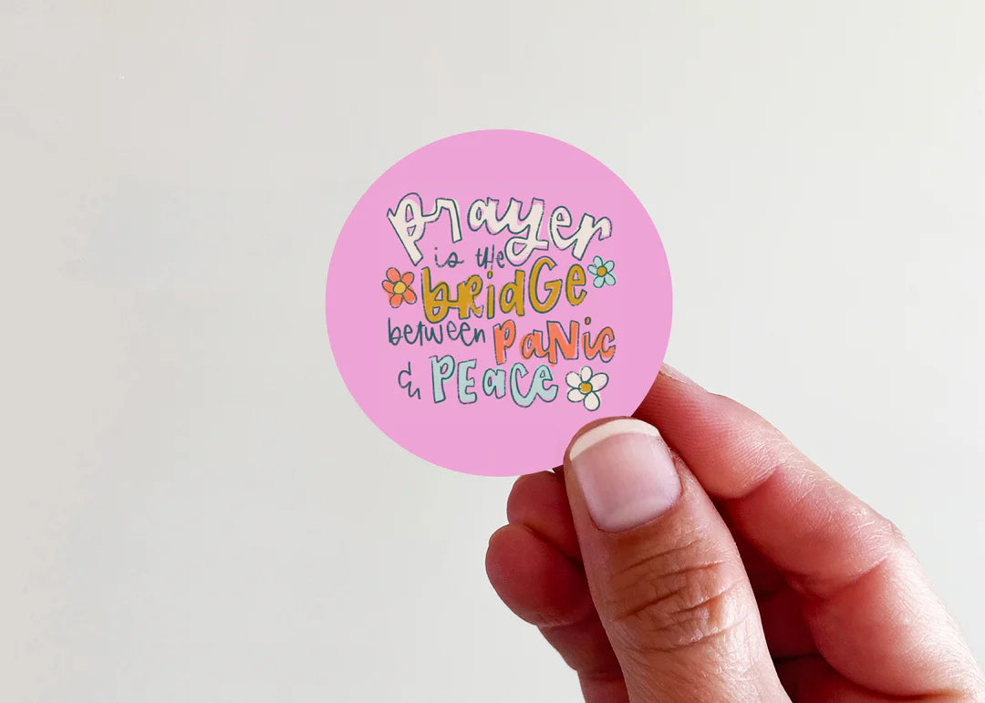 A hand holding a round pink sticker with colorful text that reads 'Prayer is the bridge between panic and peace' with small flower illustrations, against a plain light background.
