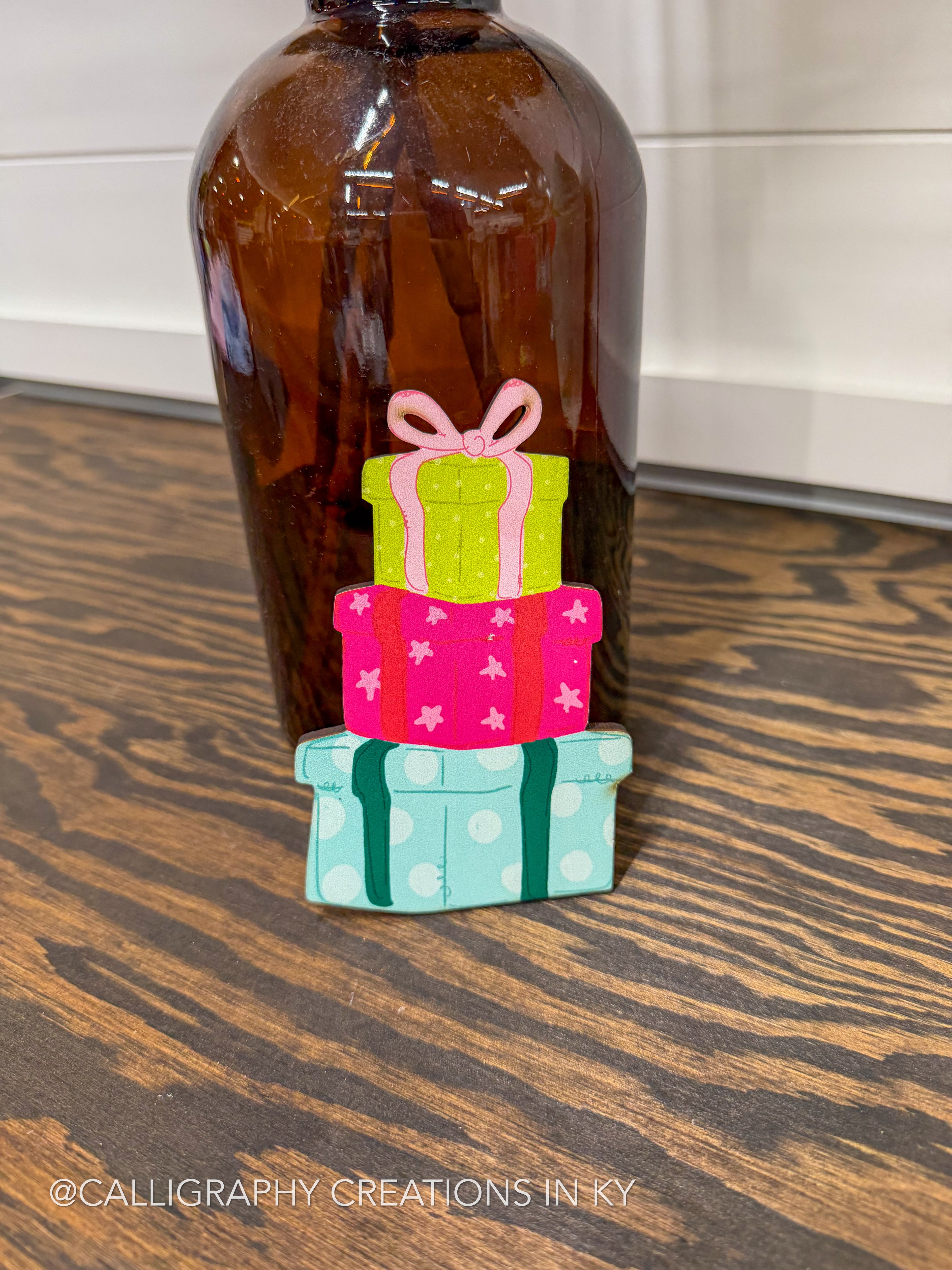 A small decorative figurine of three stacked gift boxes in bright pink, lime green, and light blue with white polka dots, placed on a dark wood tabletop in front of an amber glass bottle.