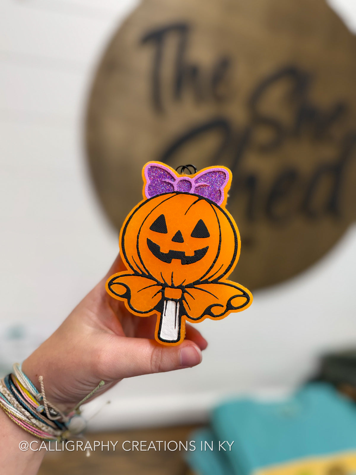 A hand holds a die-cut Halloween freshie shaped like a jack-o'-lantern lollipop with a purple glittery bow, set against a blurred background with a wooden sign.