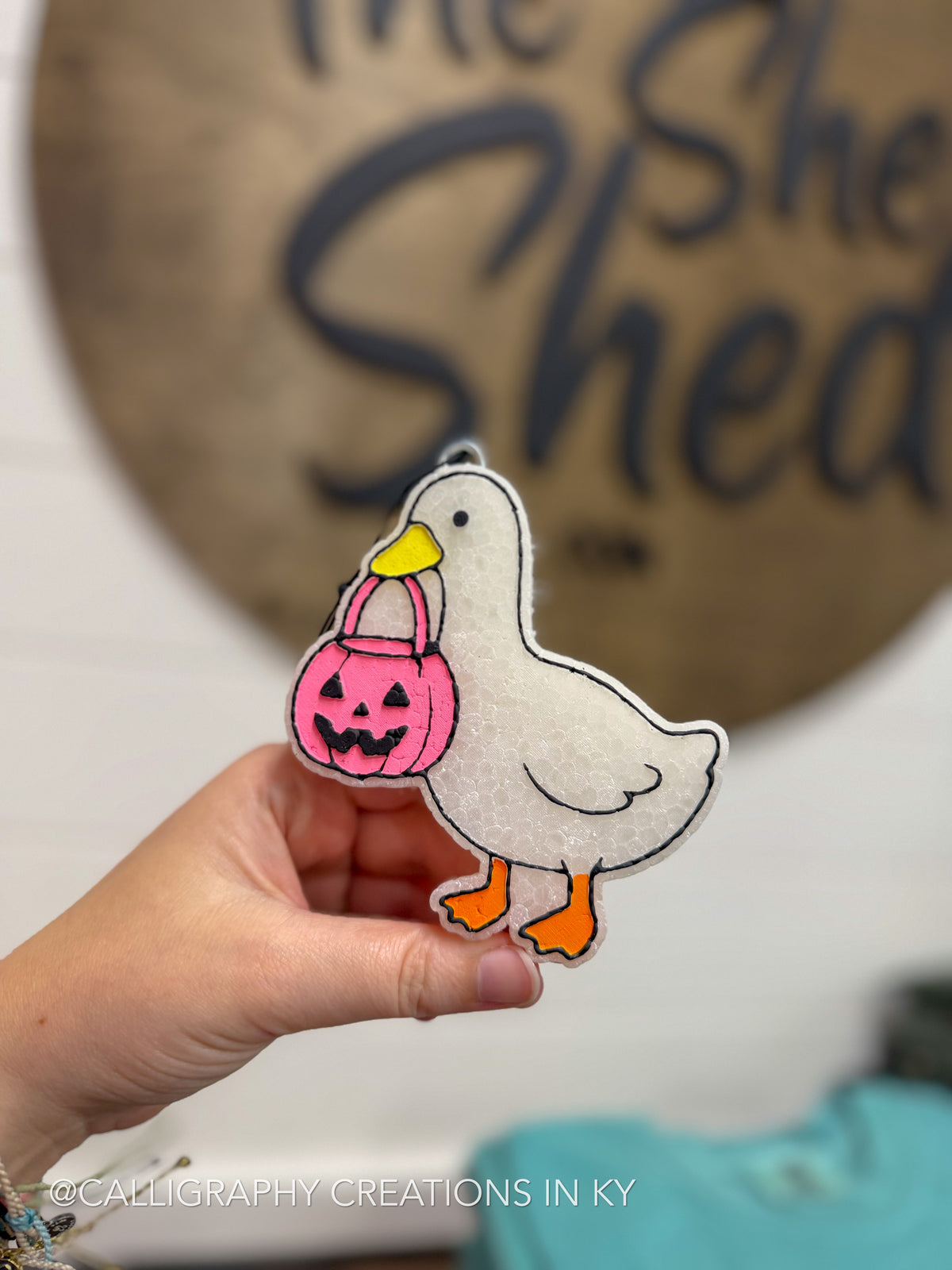 A hand holds a Pumpkin Goose Freshie Scent Sweater Weather of a white goose holding a pink pumpkin bucket, with a blurred sign in the background.