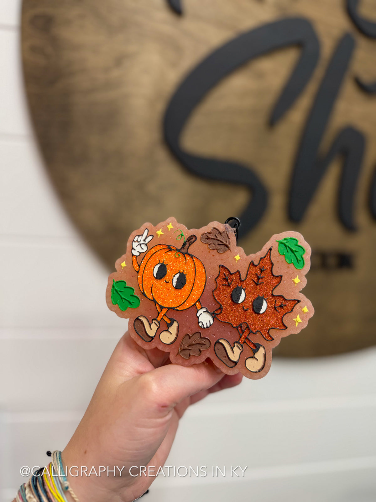 A hand holds a die-cut autumn-themed wooden freshie featuring a smiling orange pumpkin and a smiling orange leaf with small green leaves and yellow stars, set against a blurred wooden sign background.