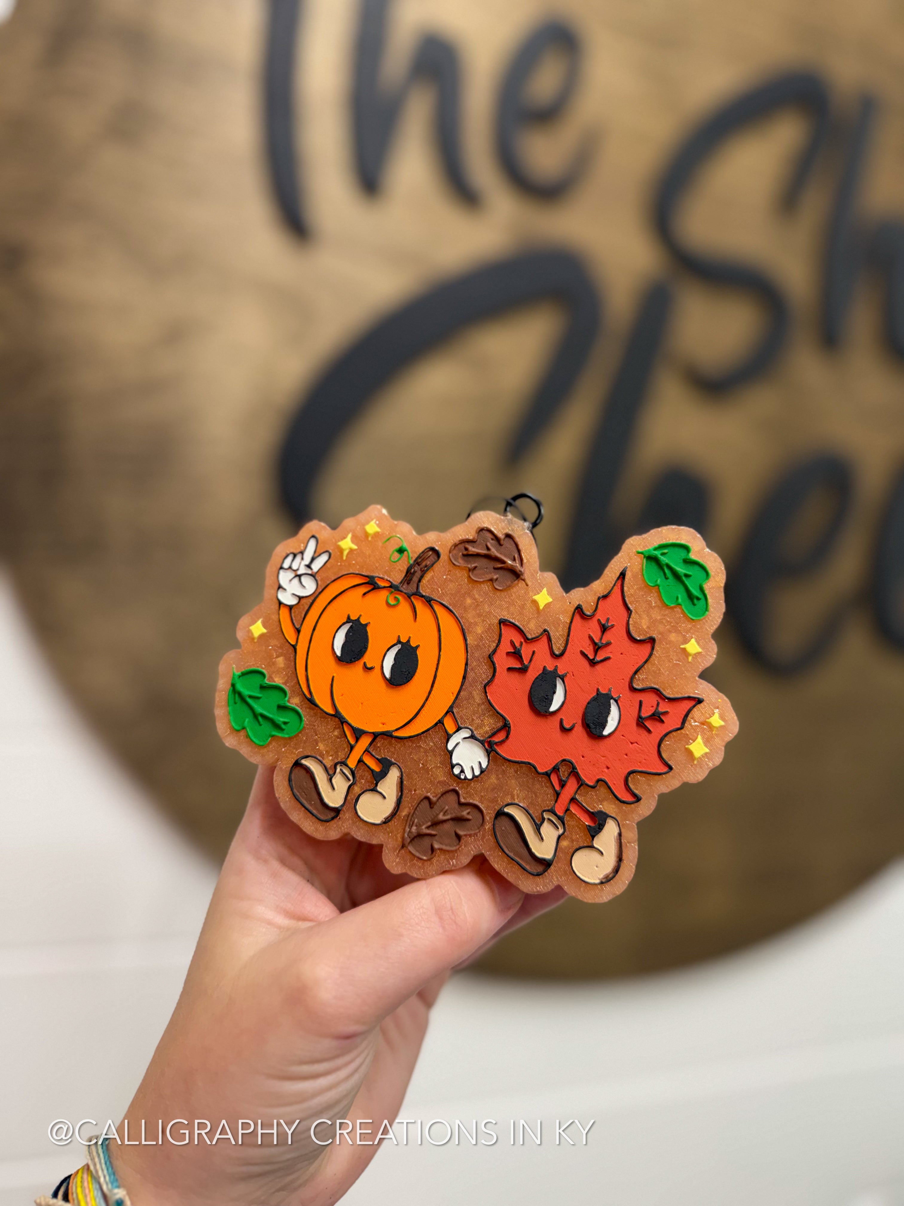 A hand holds a decorative autumn-themed freshie featuring a smiling orange pumpkin and a smiling red maple leaf character with brown acorns and green leaves, in front of a blurred wooden sign.