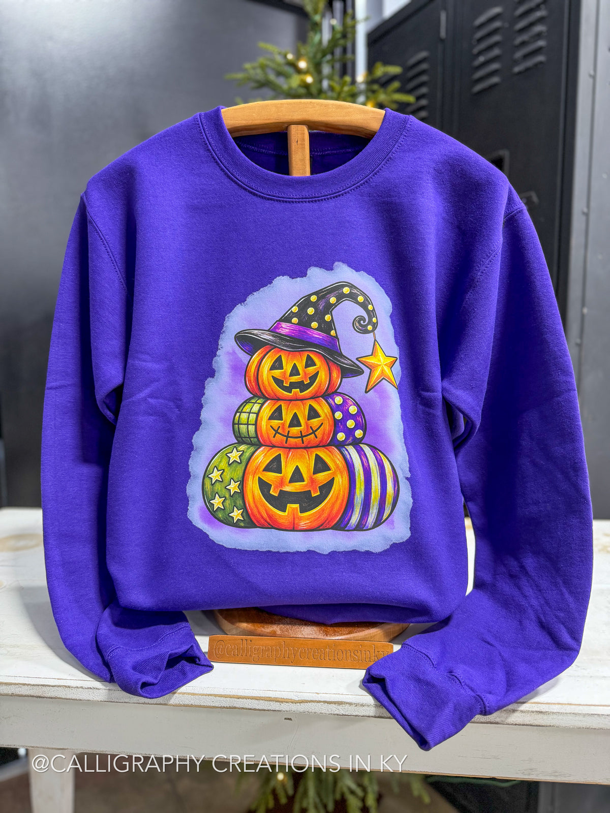 A purple crewneck sweatshirt featuring a colorful graphic of stacked jack-o'-lanterns wearing witch hats, displayed on a wooden hanger on a white table.