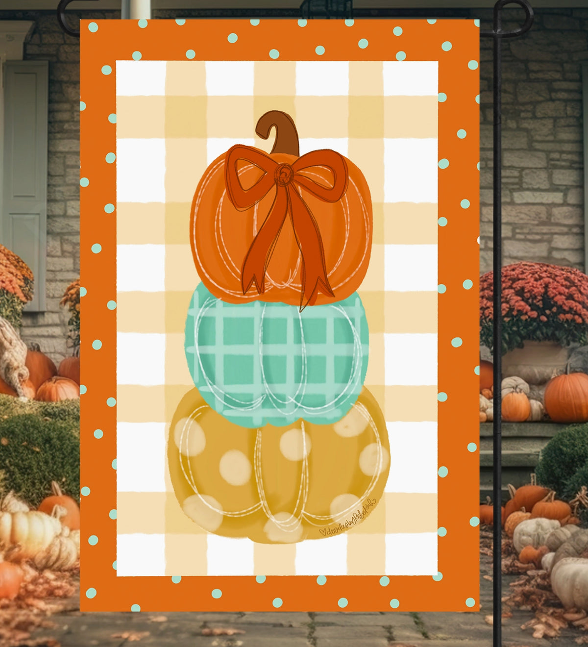 A vertical decorative garden flag displayed outdoors on a black metal stand. The flag features three stacked pumpkins in orange, mint green, and mustard yellow, set against a white and orange gingham background with a turquoise polka-dot border.
