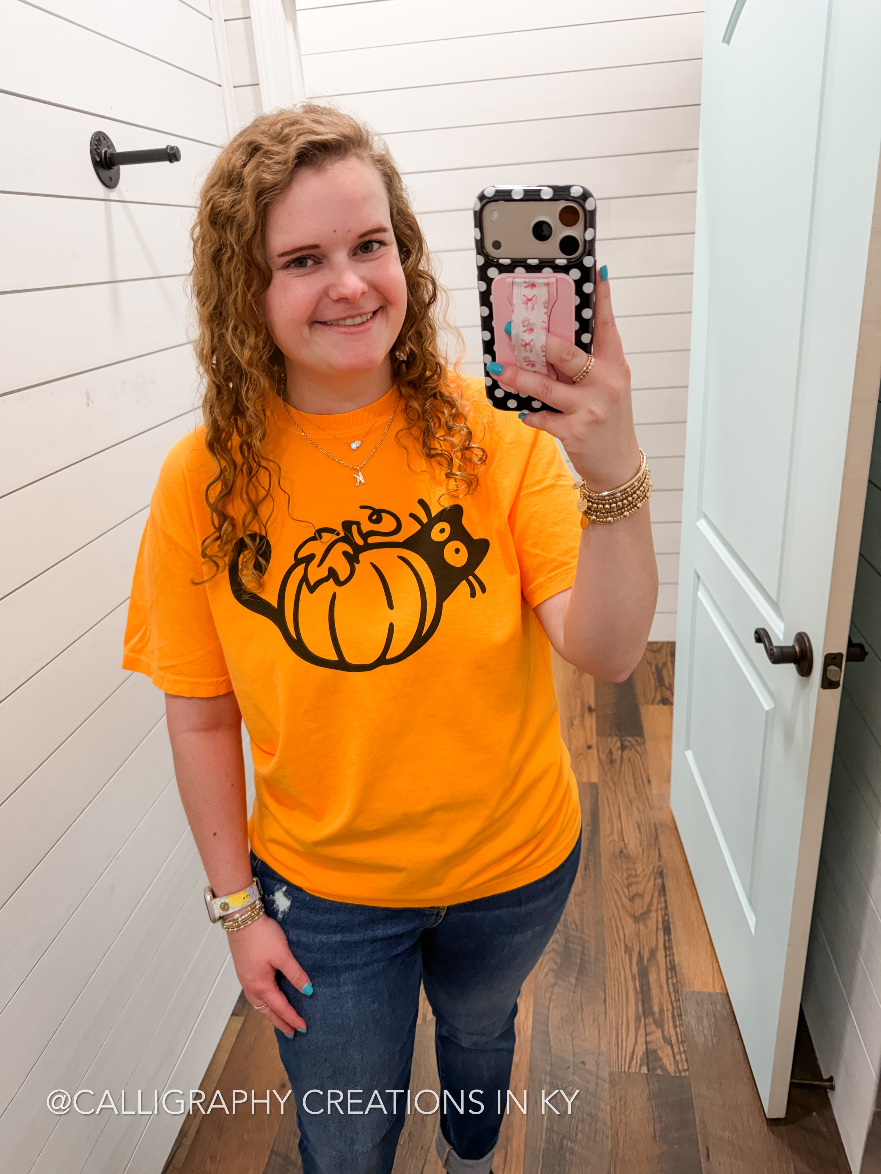 A woman takes a mirror selfie wearing a bright orange t-shirt with a black graphic of a pumpkin and cat, paired with blue jeans. The background shows a white shiplap wall and a white door.