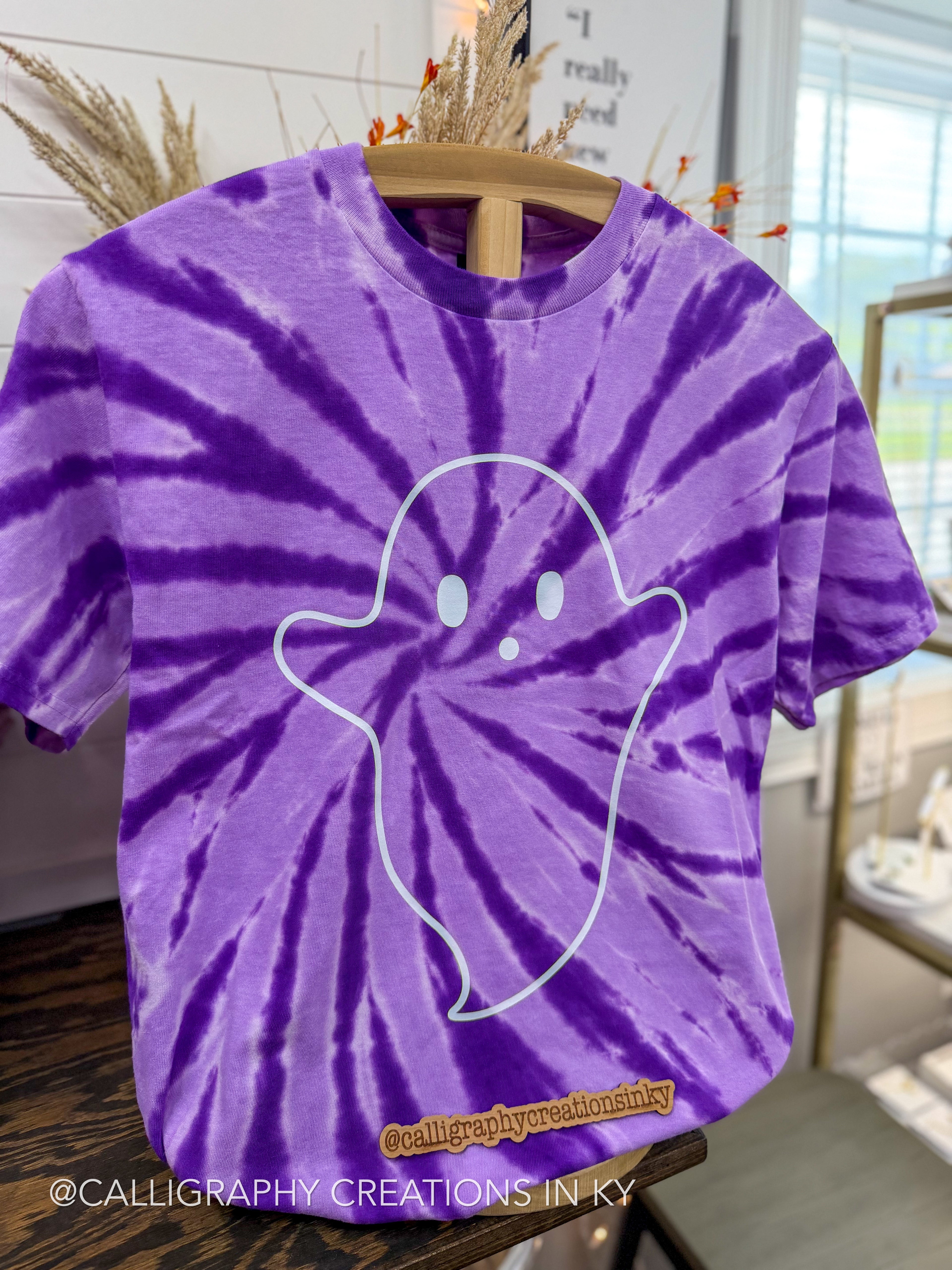 A purple and violet tie-dye t-shirt with a white ghost outline graphic on the front, hanging on a wooden hanger in a styled indoor setting.
