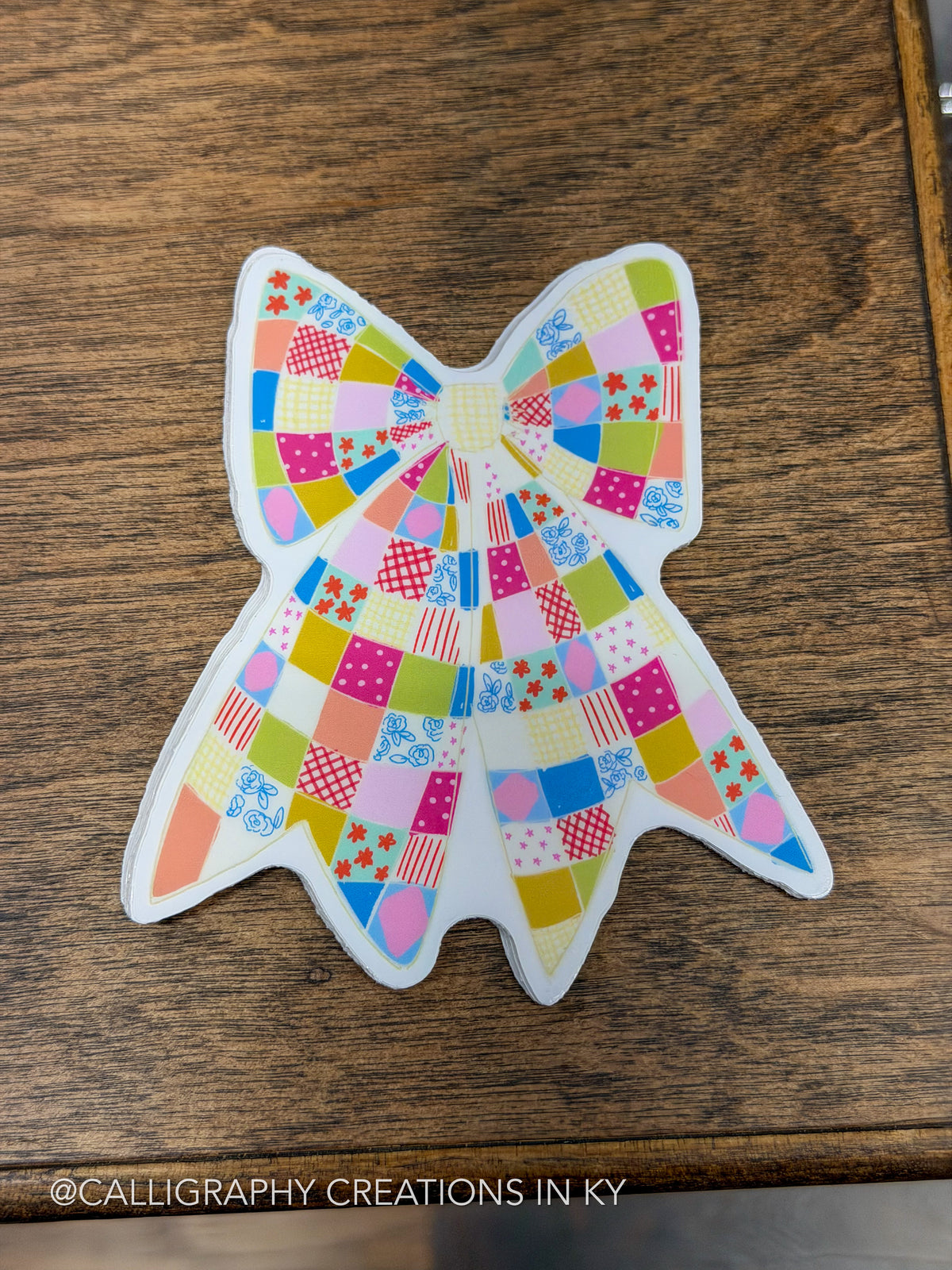 A bow-shaped patchwork sticker or decal with a colorful patchwork pattern, placed on a dark wood surface. The pattern contains various pastel and bright colors like pink, blue, yellow, green, and orange in different geometric and floral designs.