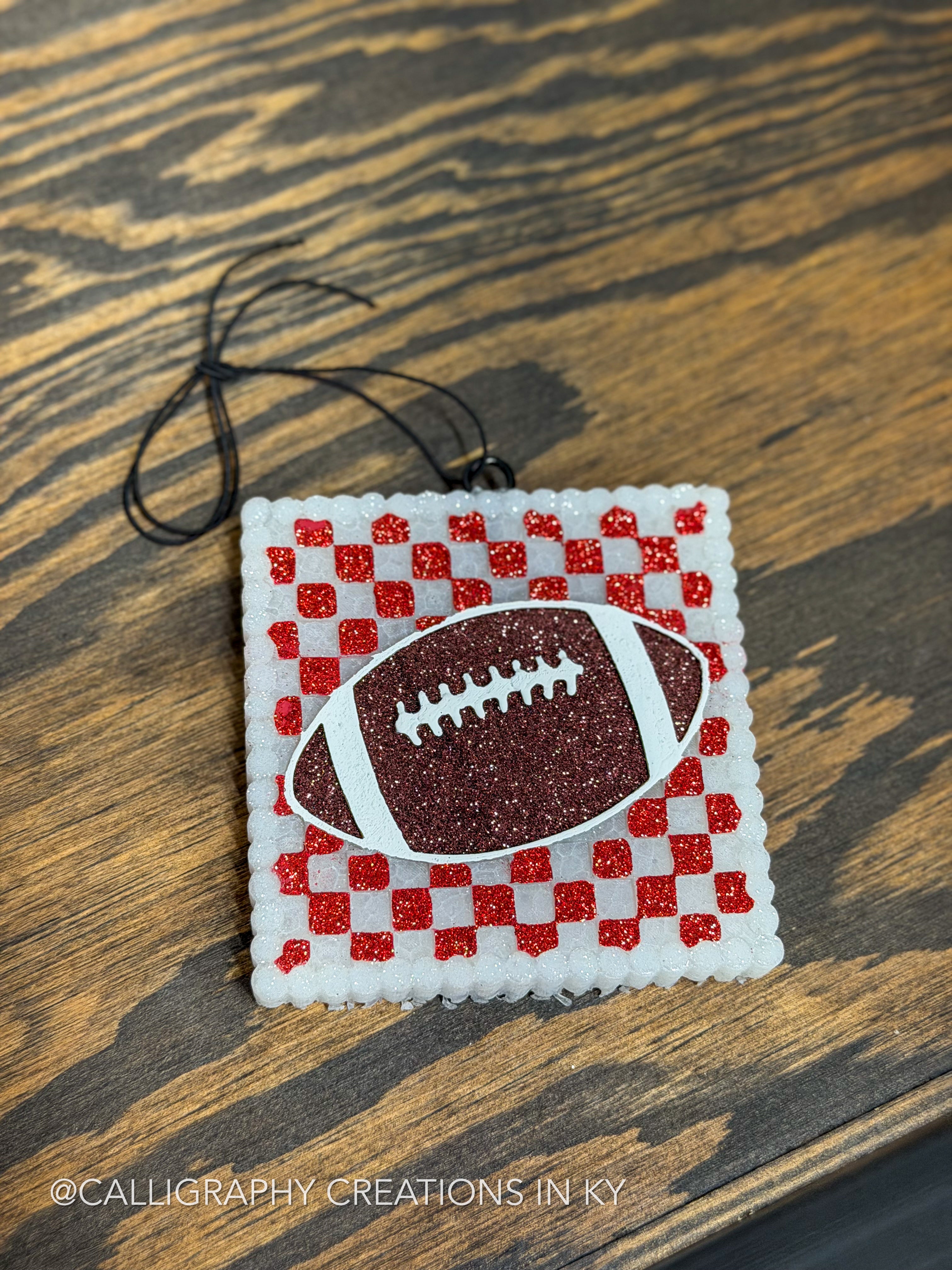 A handmade square freshie featuring a red and white checkered pattern with a glittery brown football in the center, hanging on a dark wood surface.