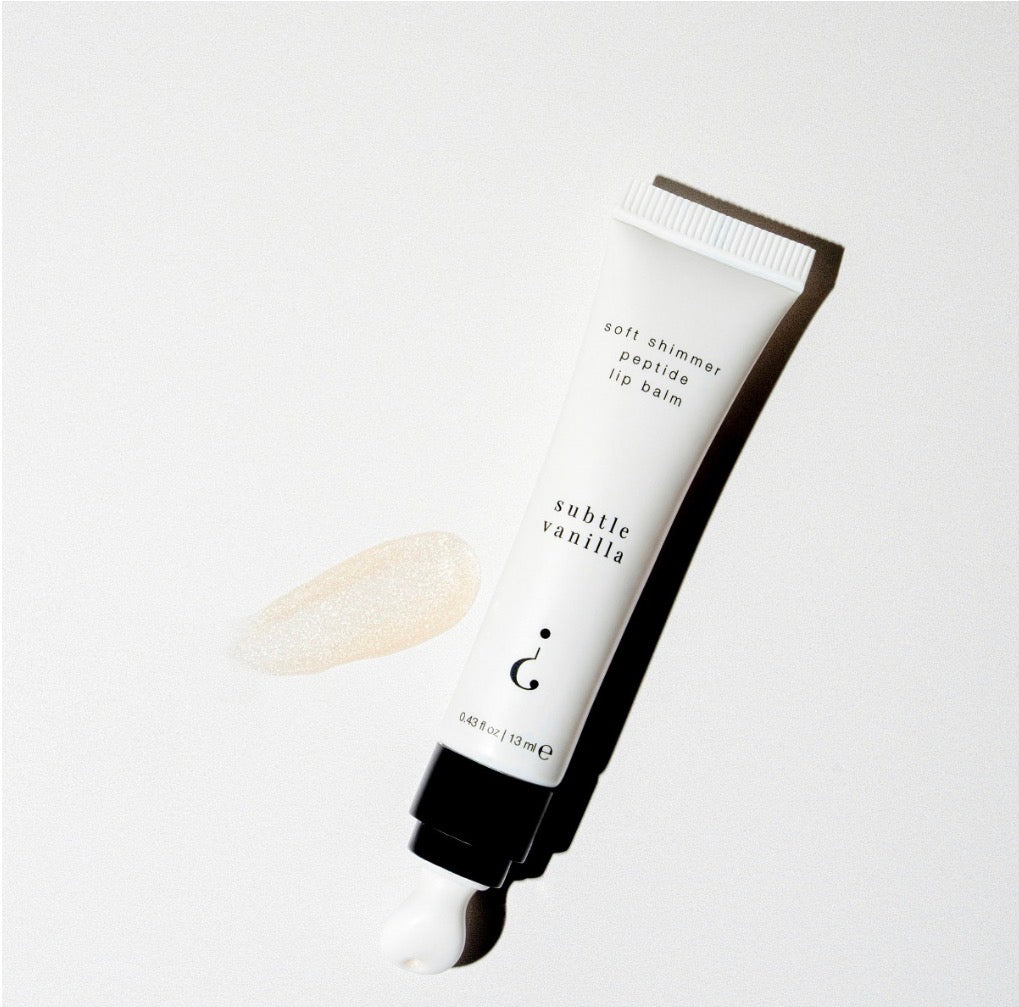 A white cosmetic tube of lip balm with black text reading 'subtle vanilla,' shown uncapped with a swatch of white balm and a black cap on a light gray background.