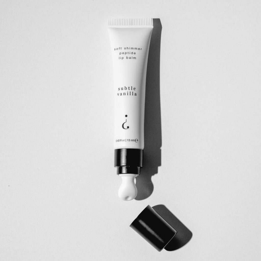 A white cosmetic tube of lip balm with black text reading 'soft shimmer peptide lip balm' and 'subtle vanilla,' shown uncapped with a dollop of white balm and a black cap on a light gray background.