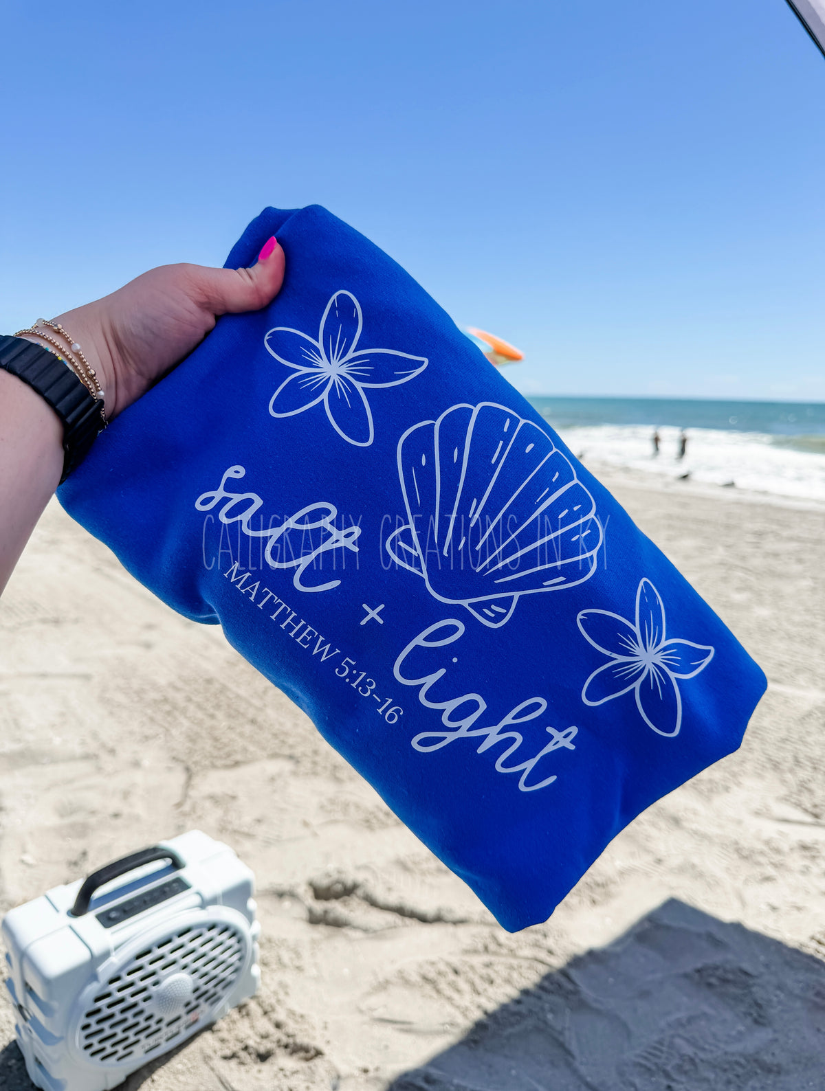 A person's hand holds a royal blue sweatshirt with white printed designs of a seashell, flowers, and the text 'salt + light' and 'MATTHEW 5:13-16' on a sandy beach with the ocean in the background.