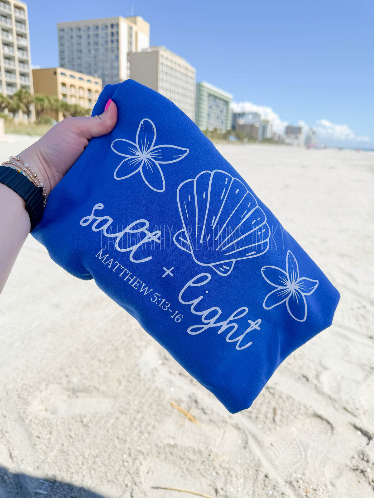 A person's hand holds a royal blue pouch or sleeve printed with white seashell and flower illustrations, along with the text 'salt + light' and 'MATTHEW 5:13-16' on a sandy beach with beachfront buildings in the background.