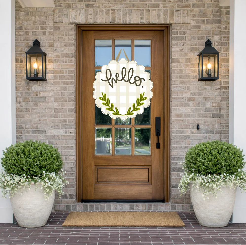 A decorative scalloped white 'hello' sign with a green leaf wreath hangs on a wooden front door in a brick entryway, flanked by two black lanterns and large potted green shrubs.