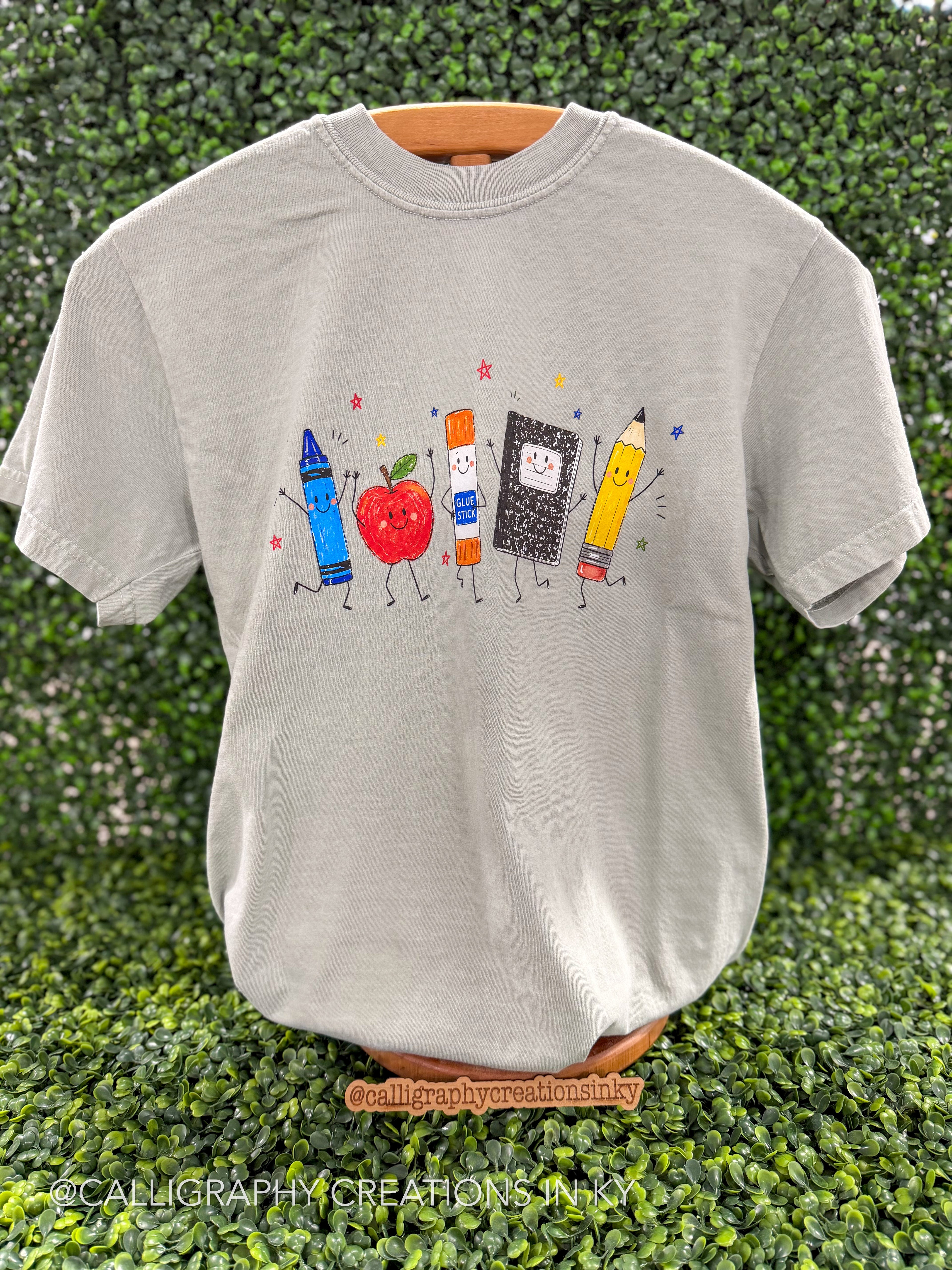 Sage green shirt with school supplies doodles on a wooden stand 