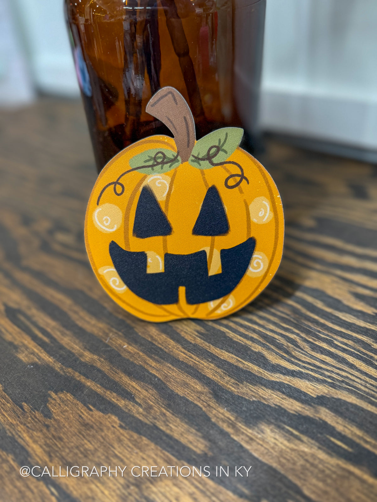 A handcrafted orange pumpkin-shaped calligraphy plaque with a black jack-o'-lantern face, green leaves, and white swirl details, displayed on a dark wood table with a blurred amber bottle in the background.