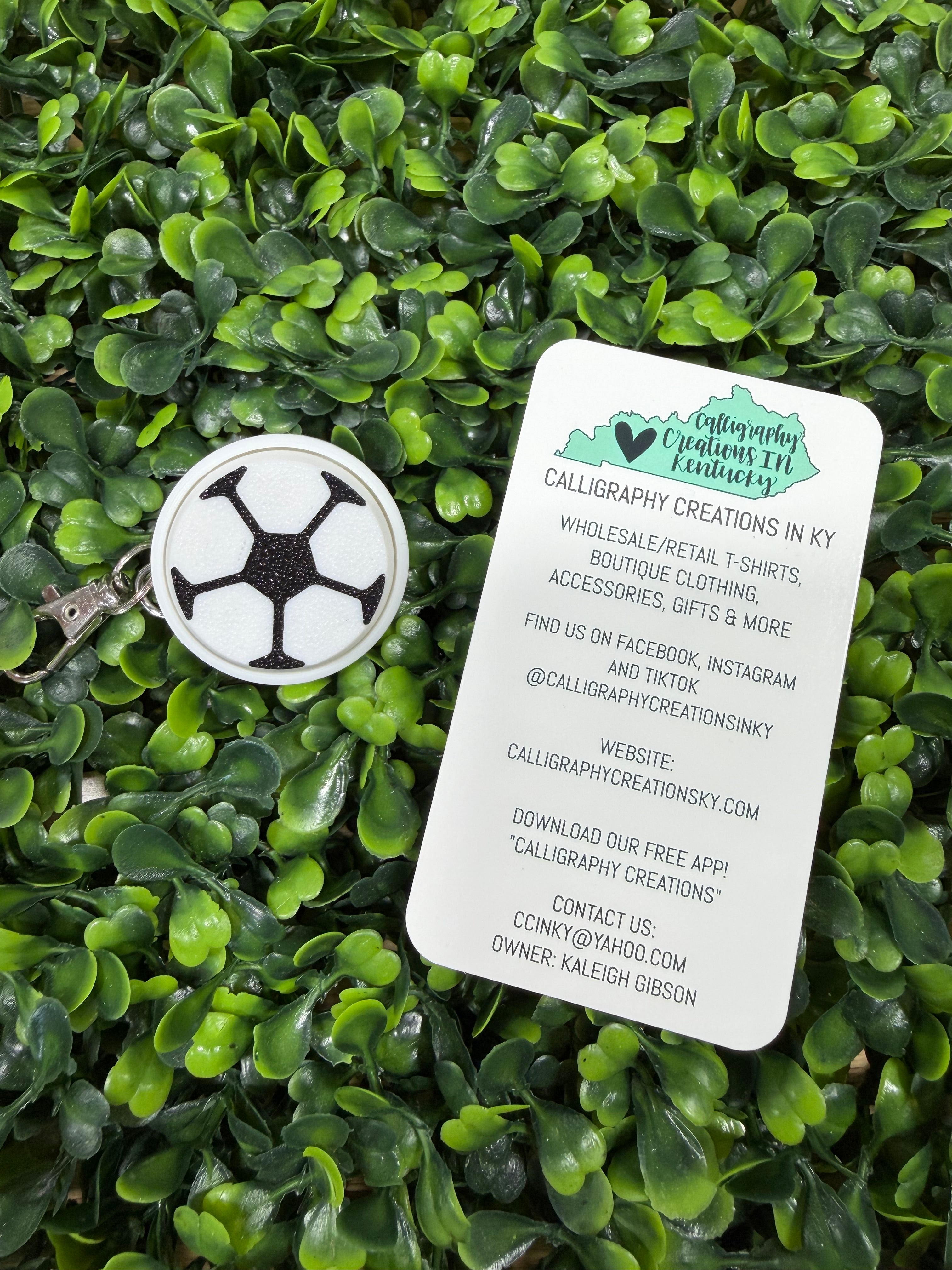A white soccer ball fidget keychain on a bed of green artificial leaves next to a white business card for Calligraphy Creations in KY.