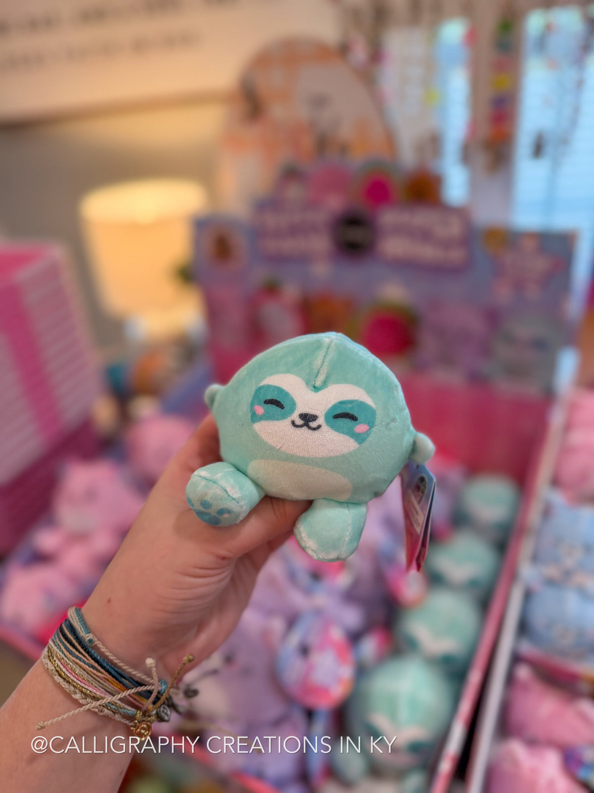 A hand holds a small plush toy shaped like a sloth in light blue and white, with a colorful, blurred toy store display in the background.